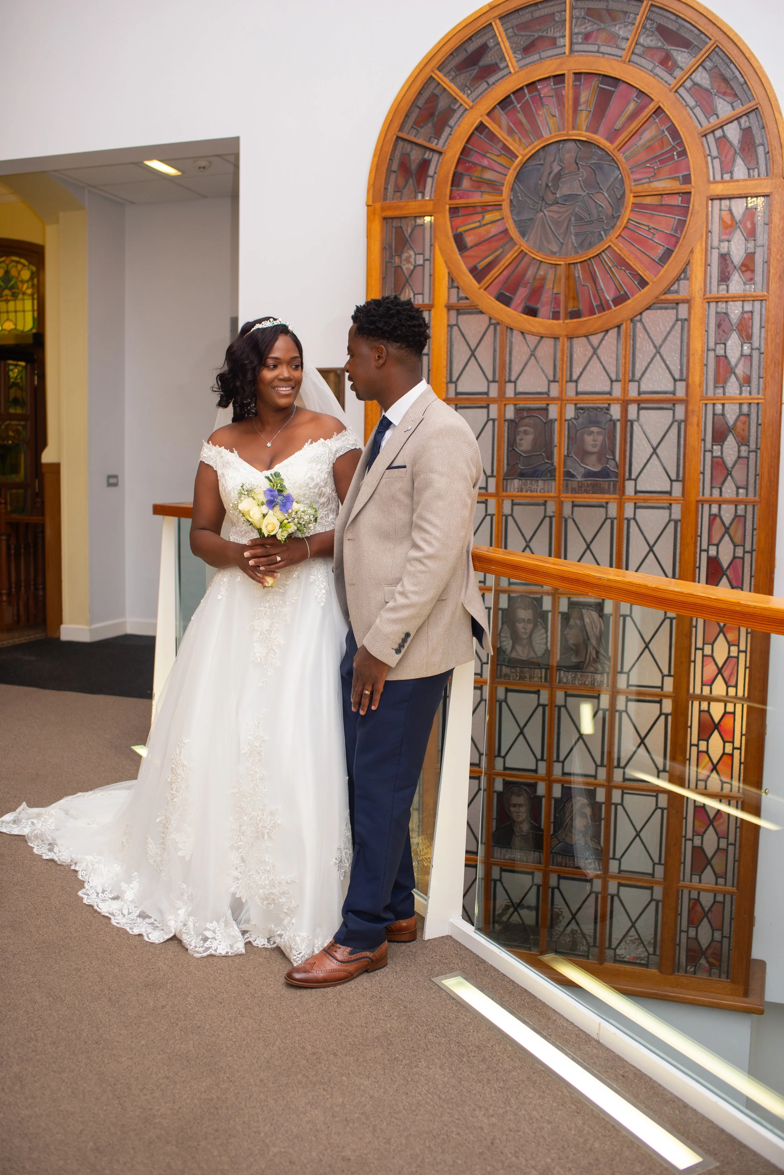 A bride and groom standing together inside a building, smiling at each other. The bride is wearing a white wedding dress with lace details, holding a bouquet of flowers. The groom is wearing a beige blazer, blue pants, and brown shoes. Behind them is