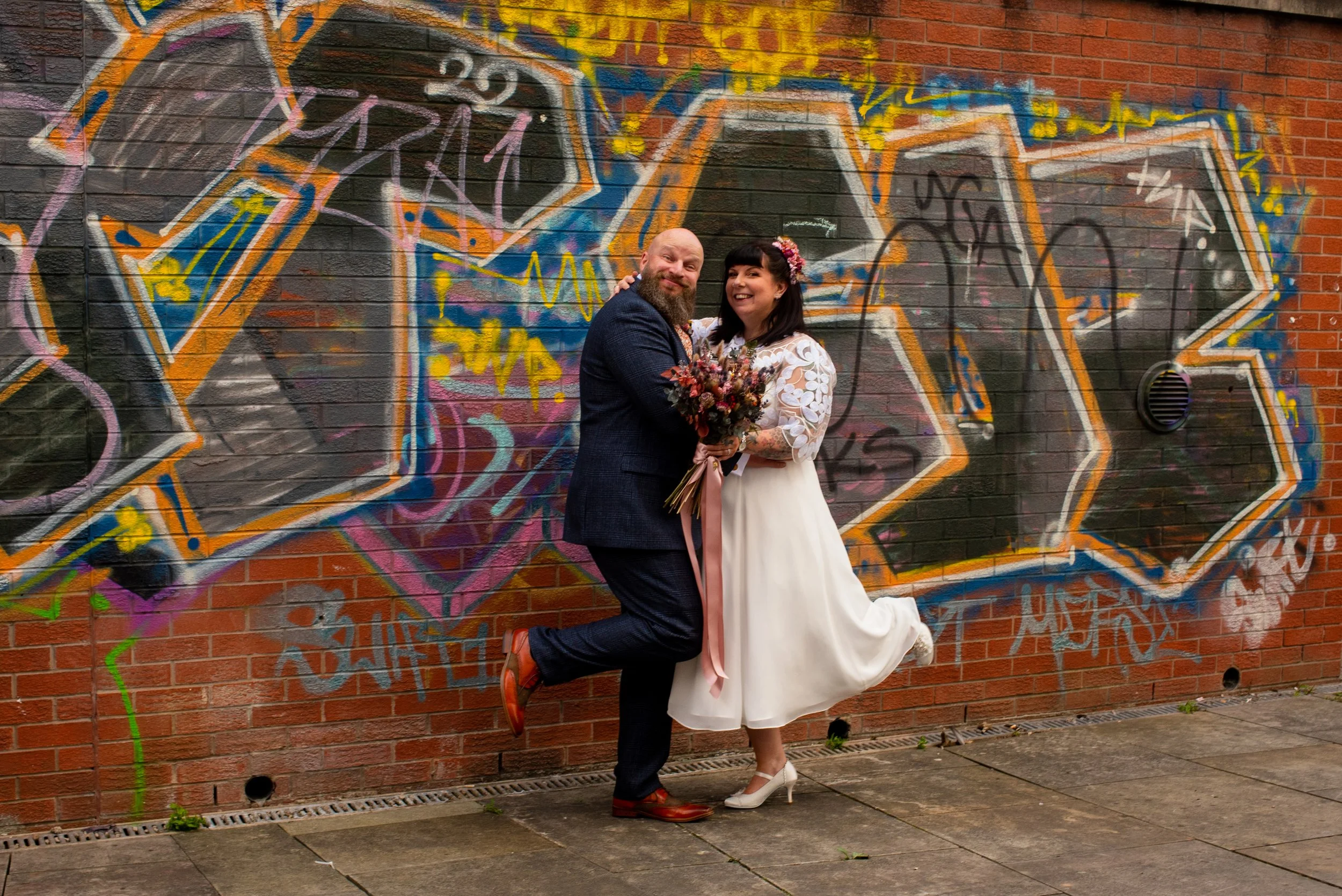 A happy couple dressed in wedding attire standing in front of a colorful graffiti wall. The man is wearing a dark suit and red shoes, holding the woman, who is in a white dress and heels, holding a bouquet of flowers.