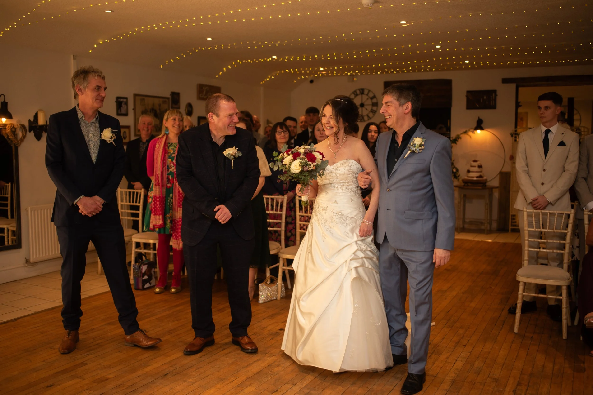 Wedding reception scene with a bride holding a bouquet, walking arm in arm with a man in a light gray suit, surrounded by guests in a warmly lit decorated venue.