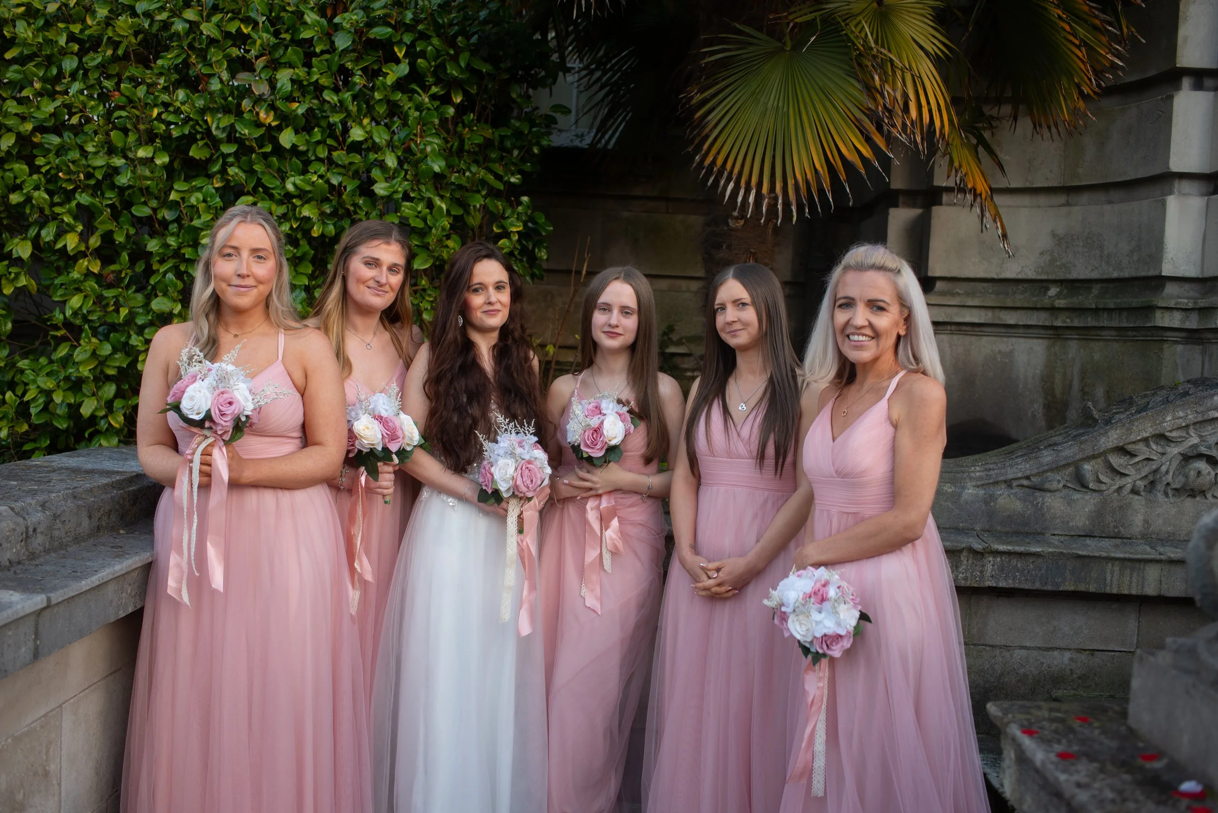 A group of six women, dressed in pink or white dresses, standing outdoors with greenery and a stone wall in the background, holding bouquets of flowers, likely at a wedding.