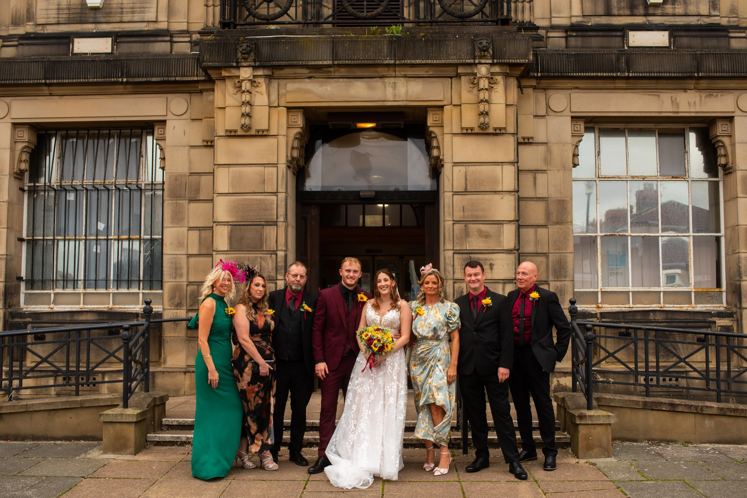 Group of people standing in front of an old stone building during a wedding. The bride is holding a bouquet of flowers, surrounded by friends and family dressed in colorful formal attire.