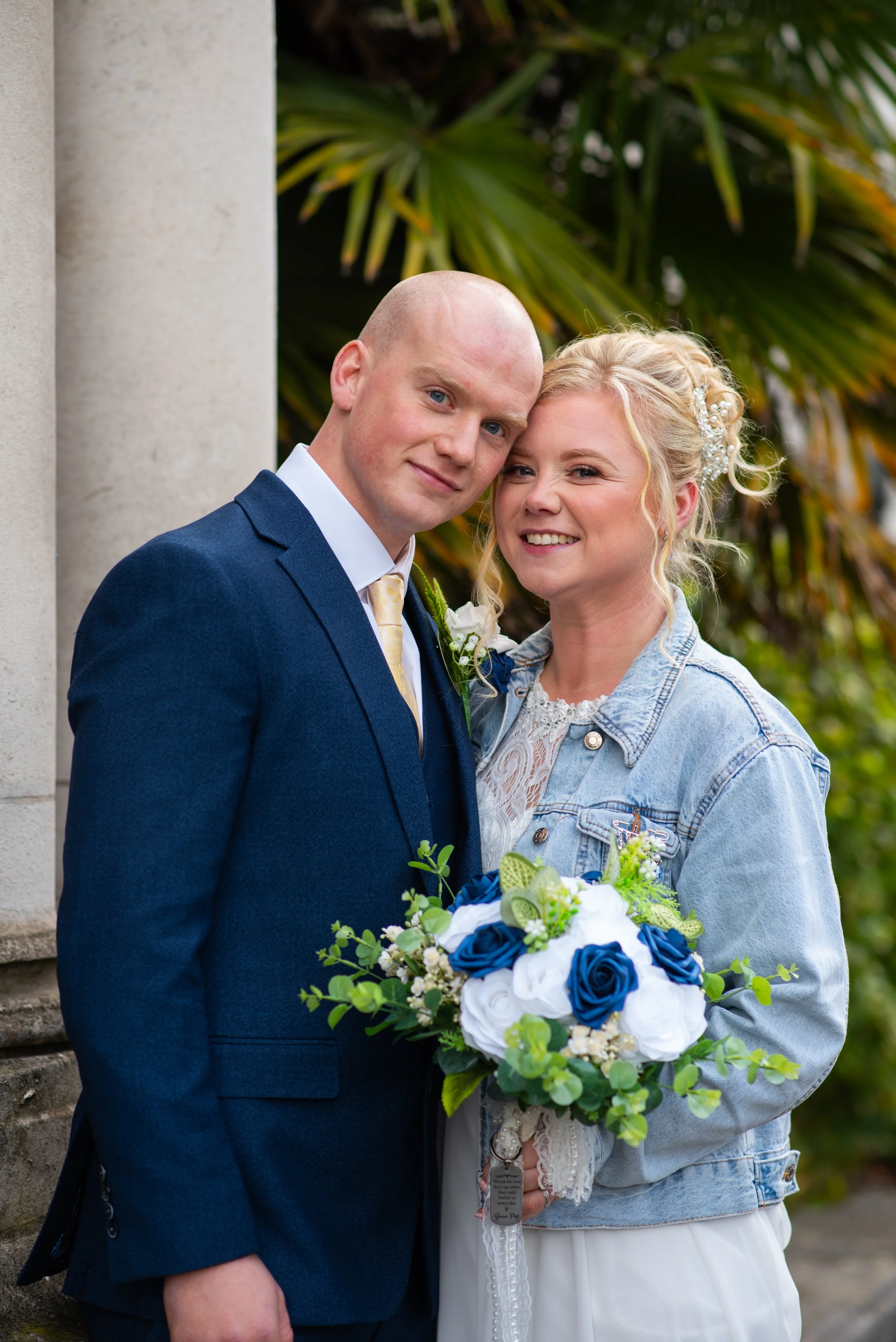 A bride and groom posing together outdoors, with the bride holding a bouquet of blue and white roses and greenery, dressed in wedding attire, after a wedding ceremony.