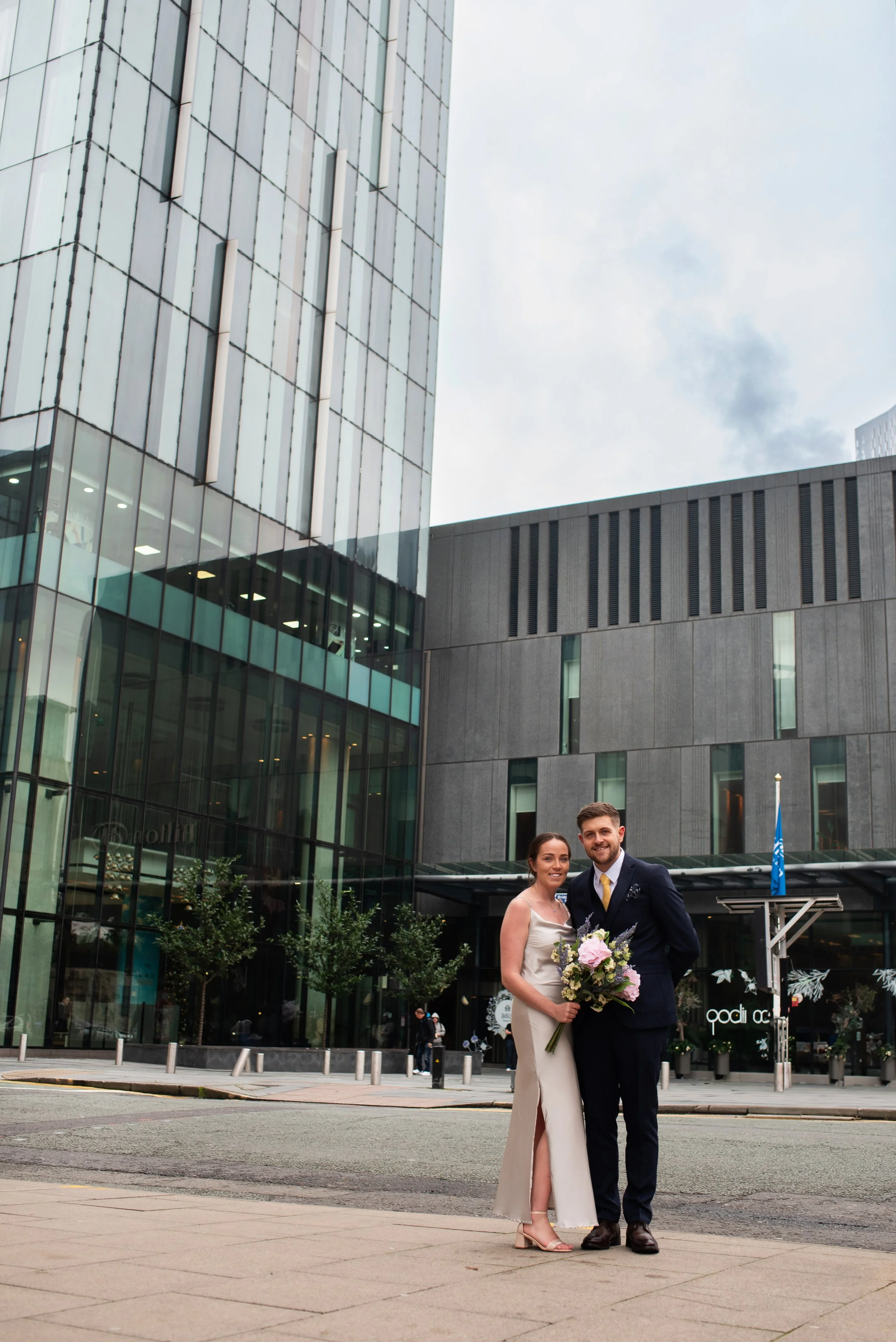 A newly married couple standing outside a modern glass building, with the bride holding a bouquet of flowers and both smiling.
