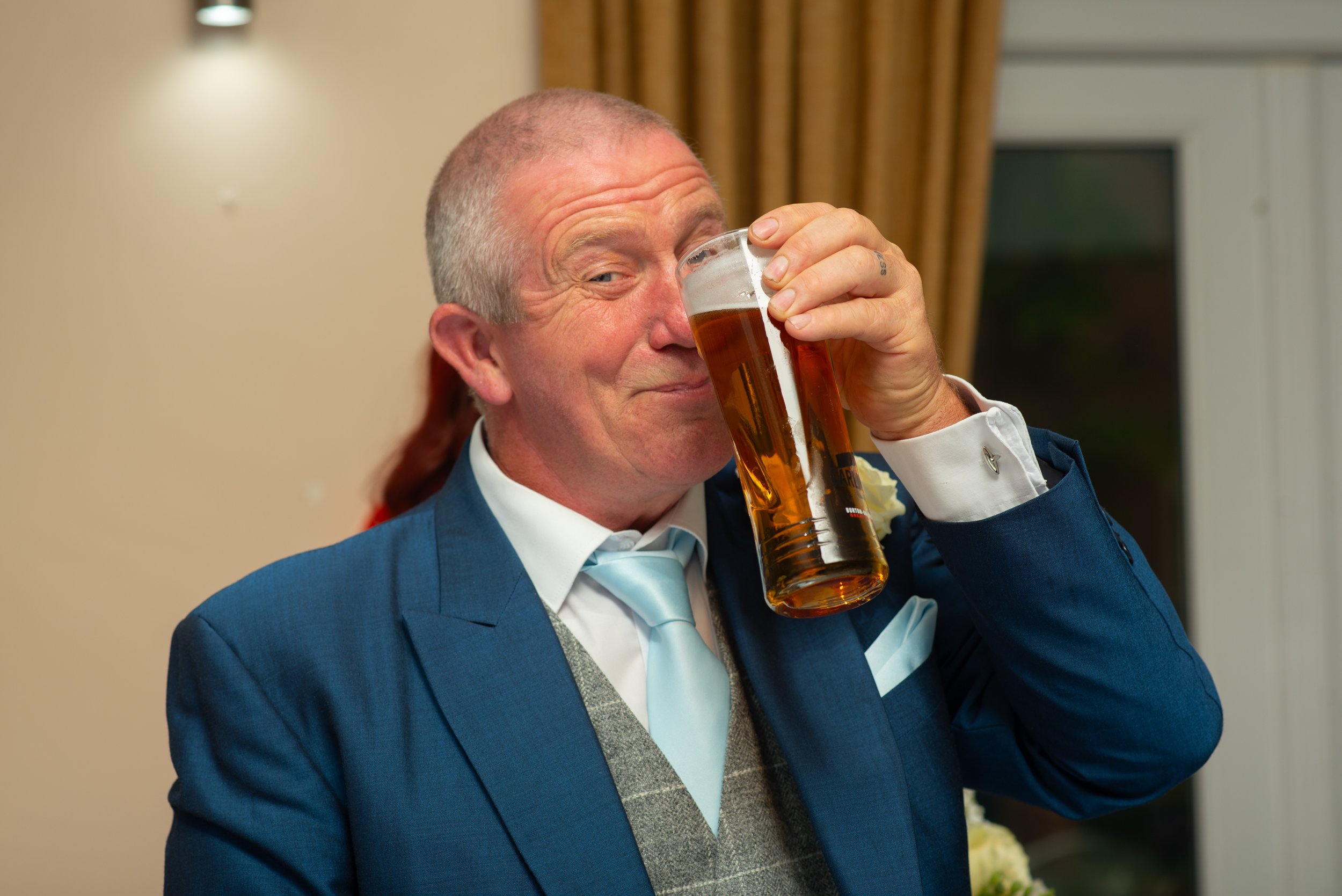 Man in a blue suit drinking beer from a tall glass, smiling with his eyes closed.