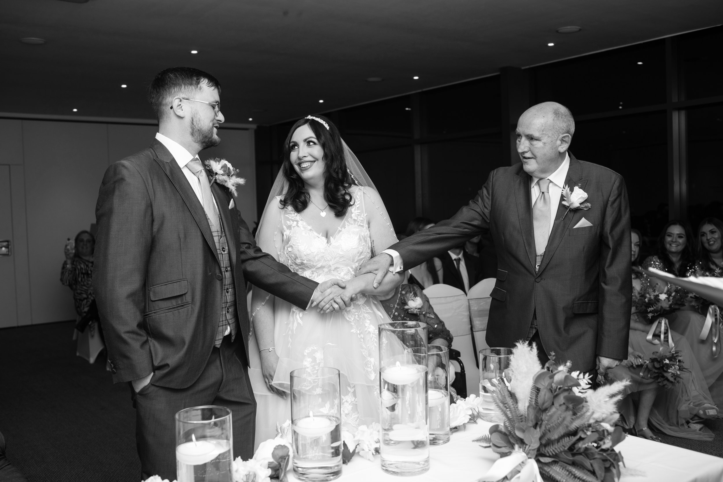 A bride and groom hold hands during a wedding ceremony, shaking hands with an officiant in a formal setting with seated guests and floral decorations.