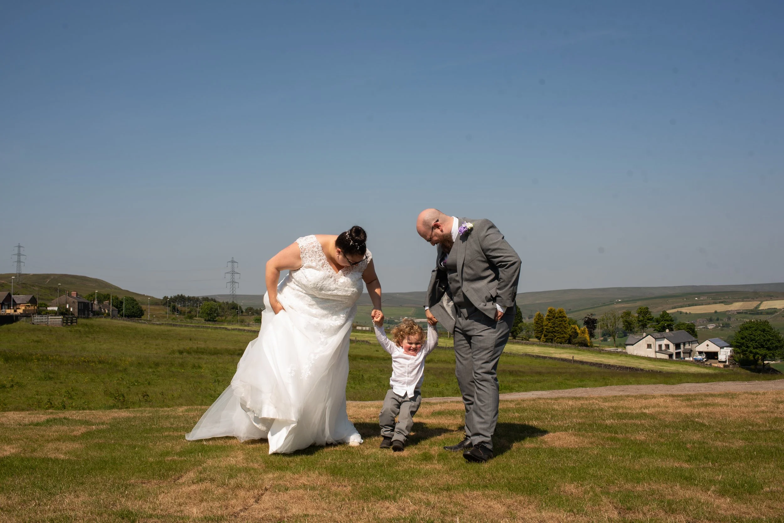 A wedding scene outdoors with a bride, groom, and a young child on a grassy field under a clear blue sky.