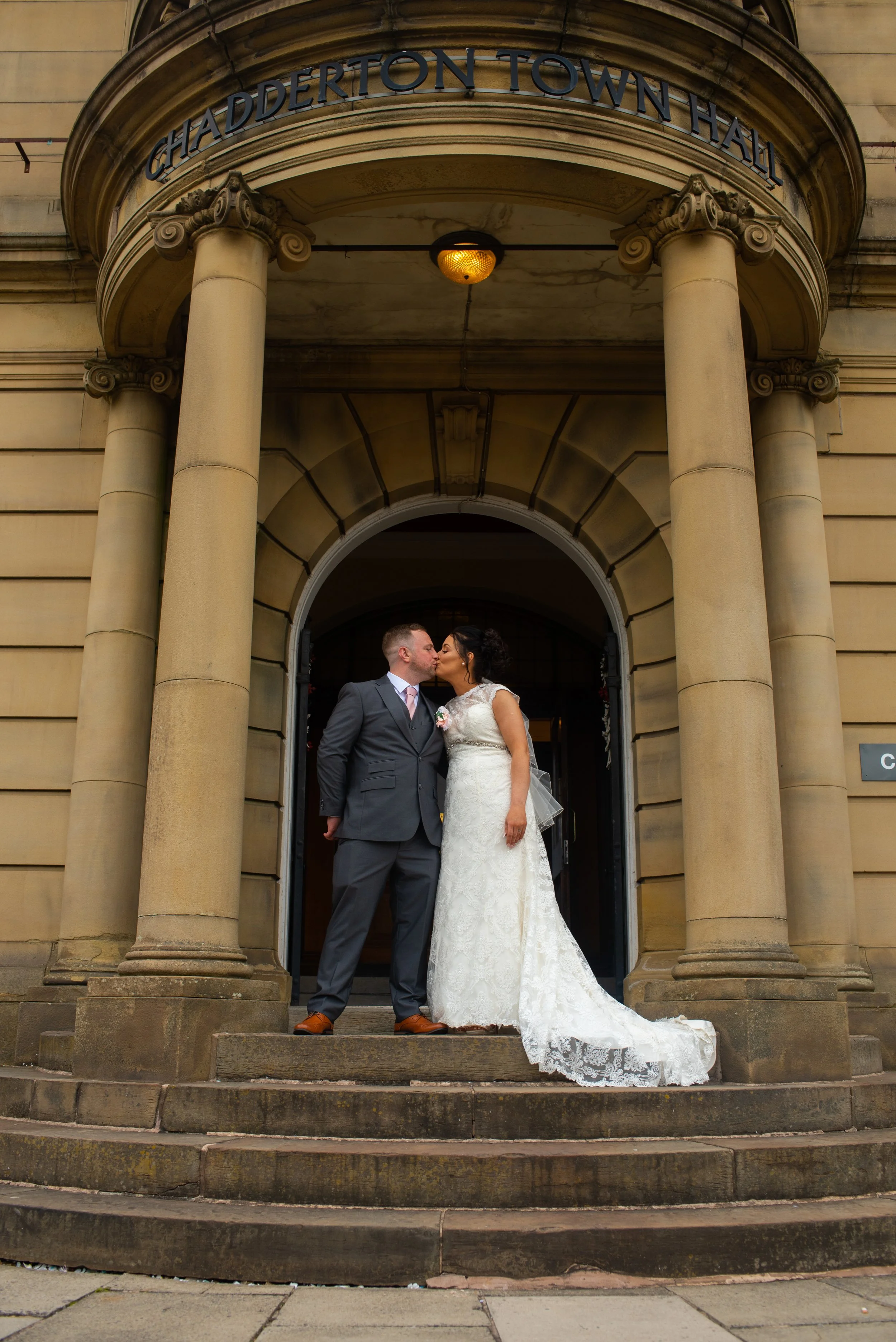A newlywed couple kissing on the steps in front of Chadderton Town Hall, with the bride wearing a white lace wedding dress and the groom in a gray suit.
