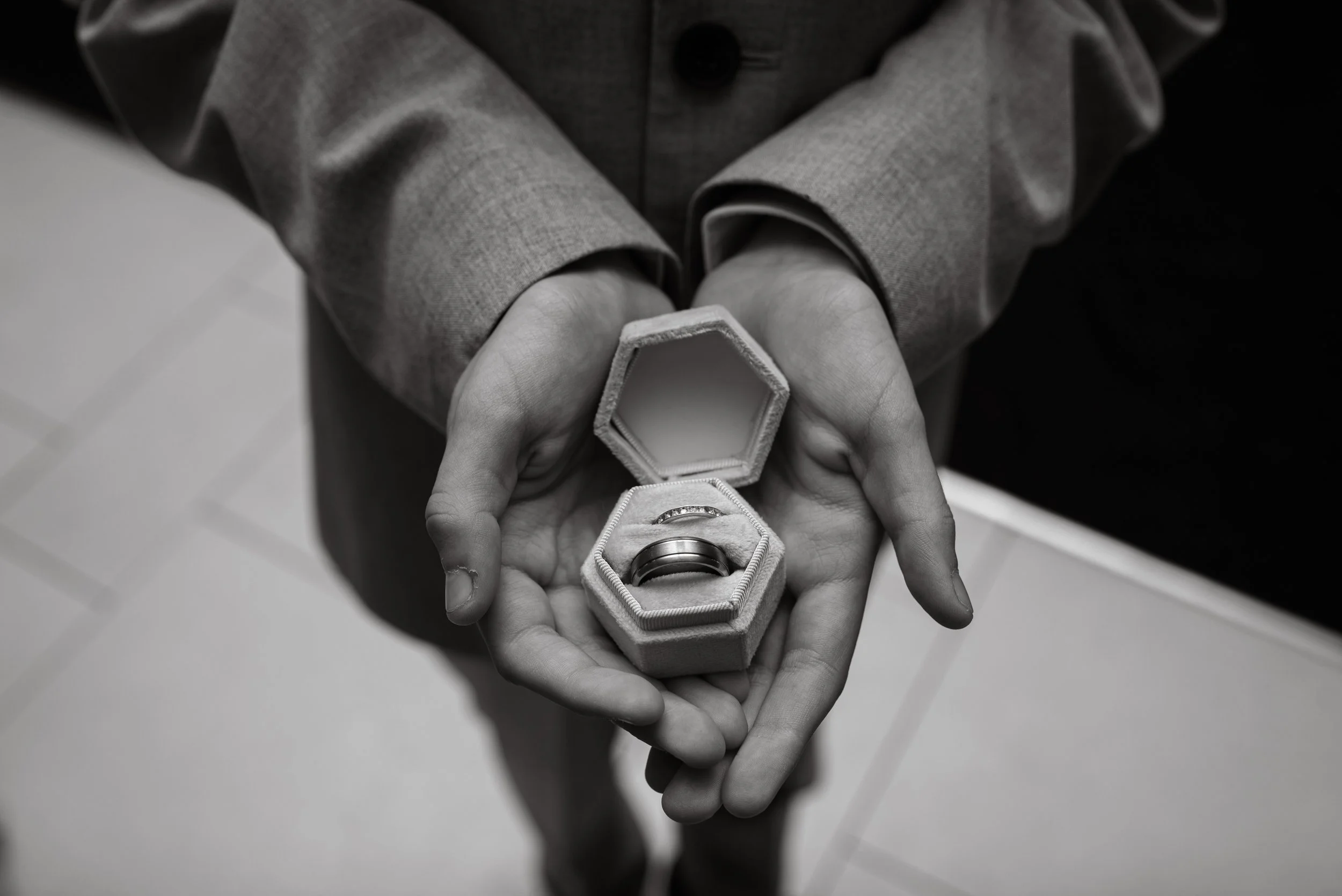 Person holding open jewelry box with wedding rings inside, with a tiled floor in the background.