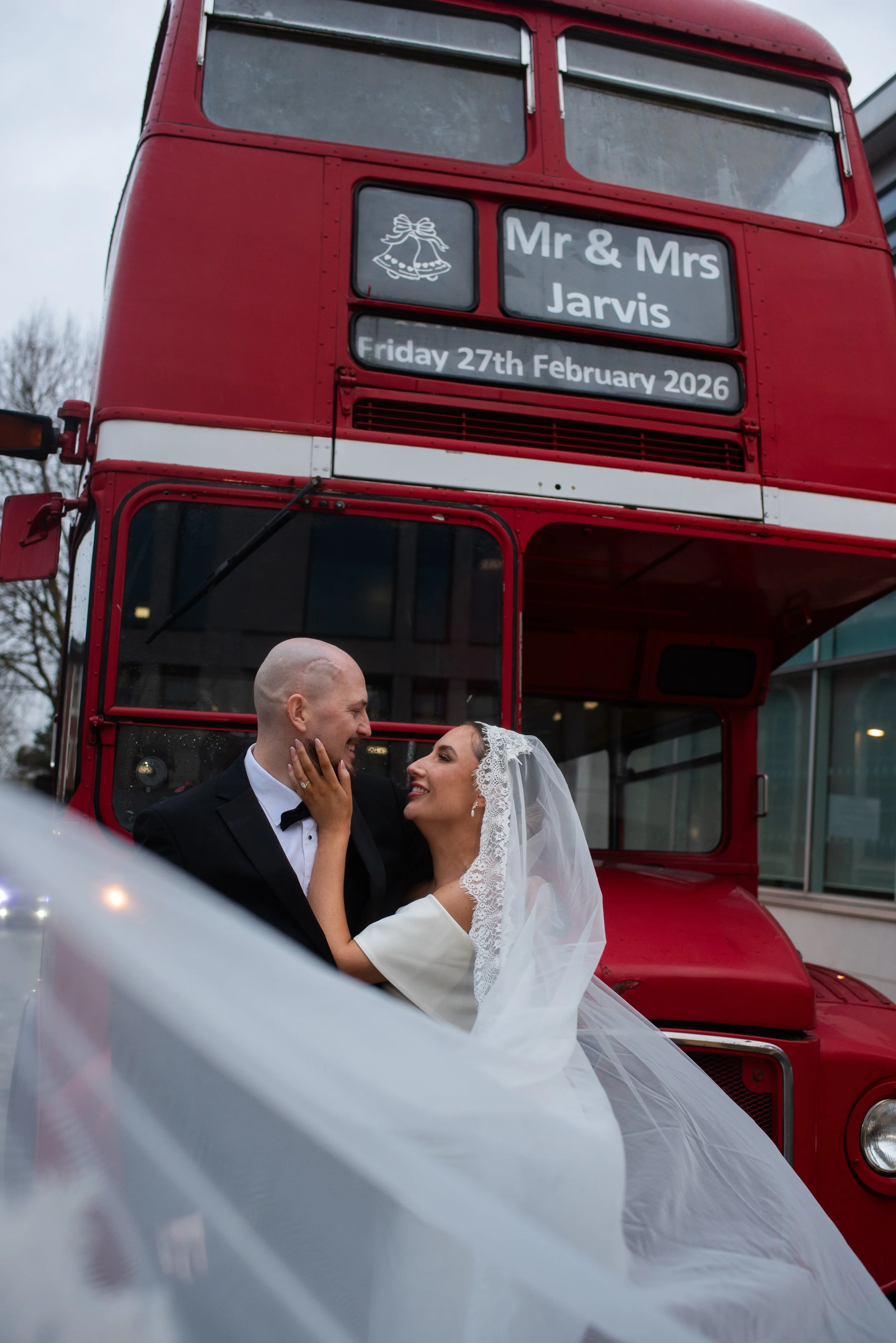 A bride and groom in wedding attire sharing a joyful moment in front of a red double-decker bus with a sign displaying the date, February 27th, 2026, and the names Mr. & Mrs. Jarvis.