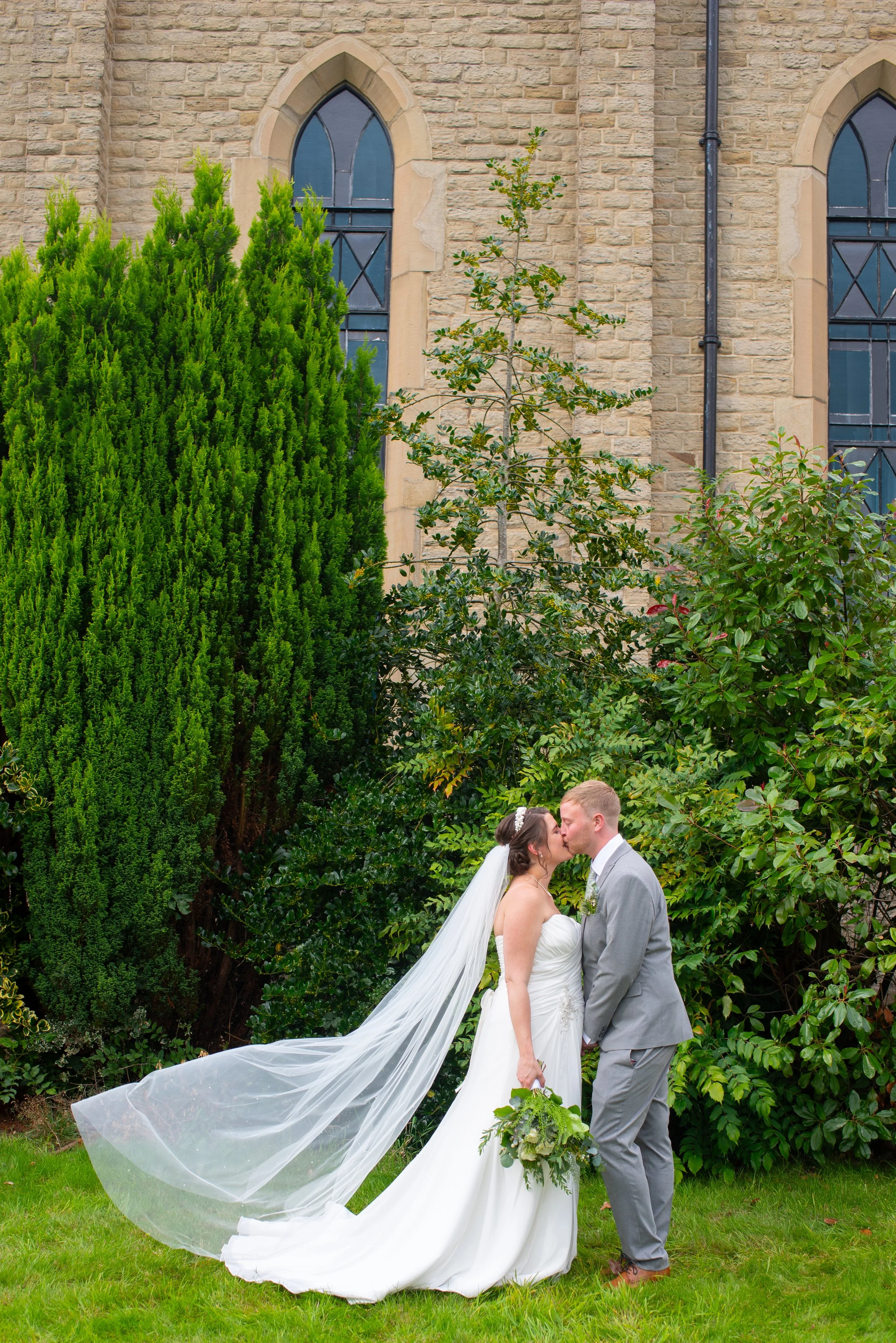 A bride in a white wedding dress and veil and a groom in a gray suit are sharing a kiss outdoors, surrounded by lush green bushes and a stone church with arched windows in the background.