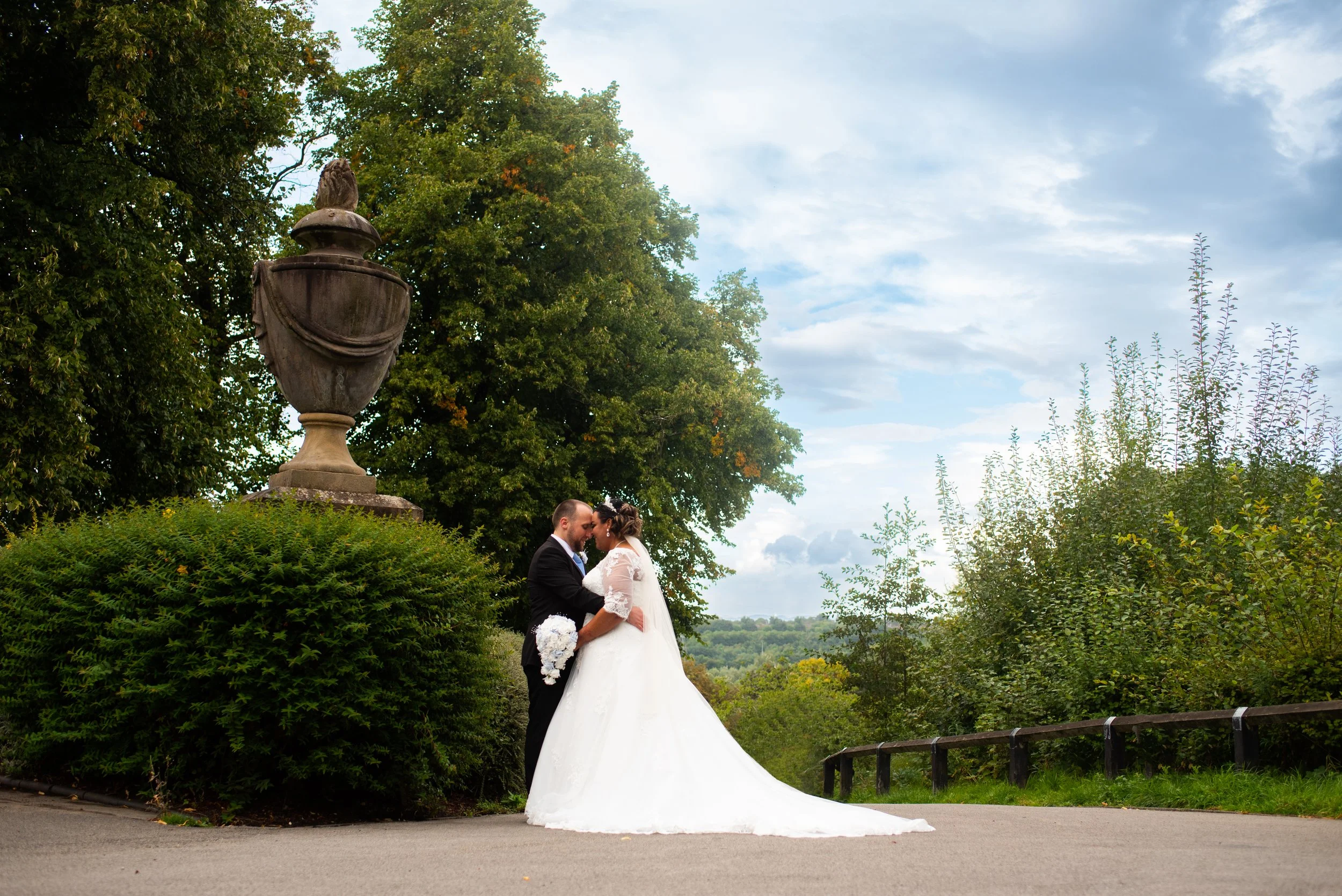 A bride and groom in wedding attire sharing a kiss outdoors, with greenery and a large decorative urn on a pedestal behind them.