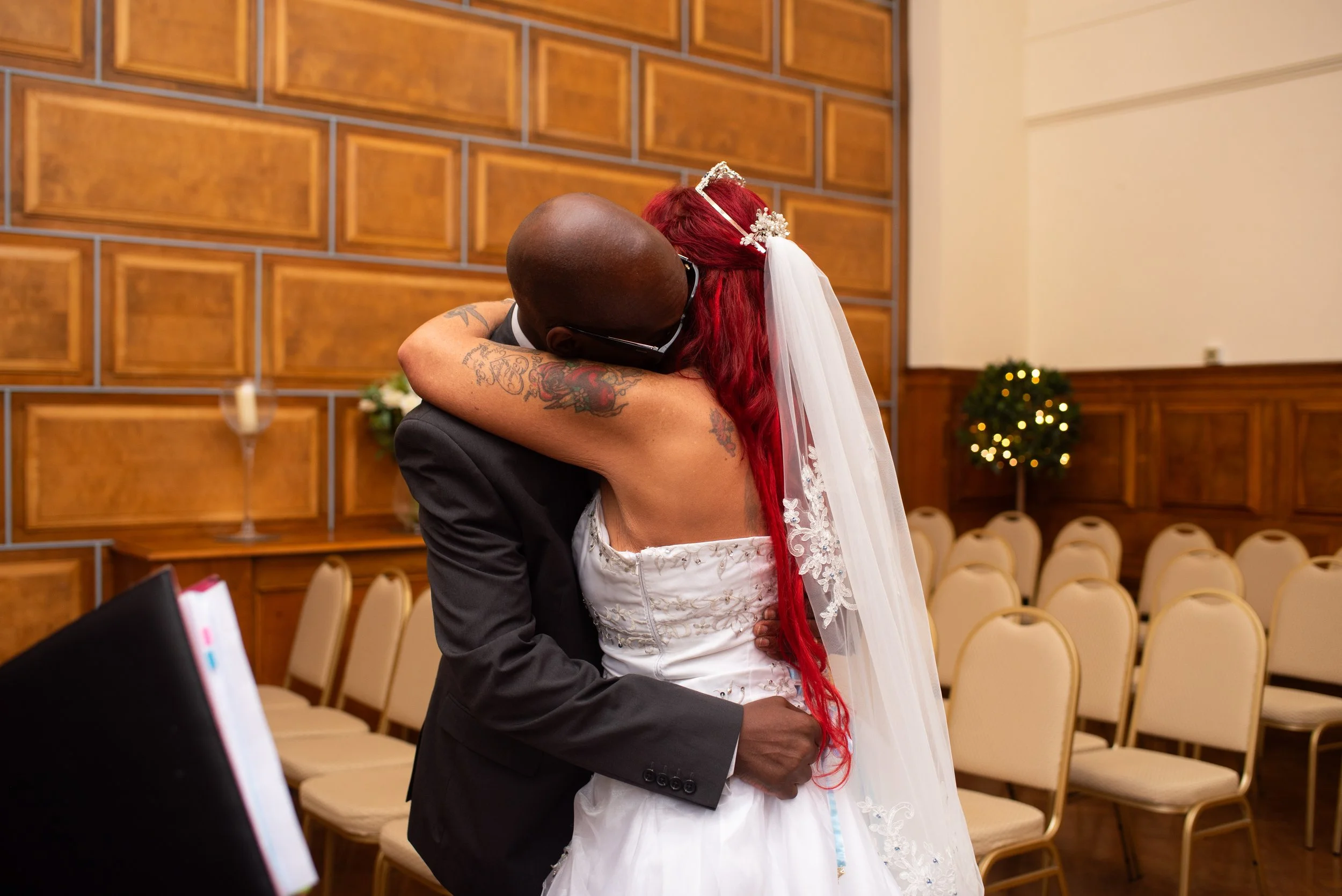 A bride with red hair and tattoos embracing a groom in a suit inside a decorated room with wooden panels and empty chairs.