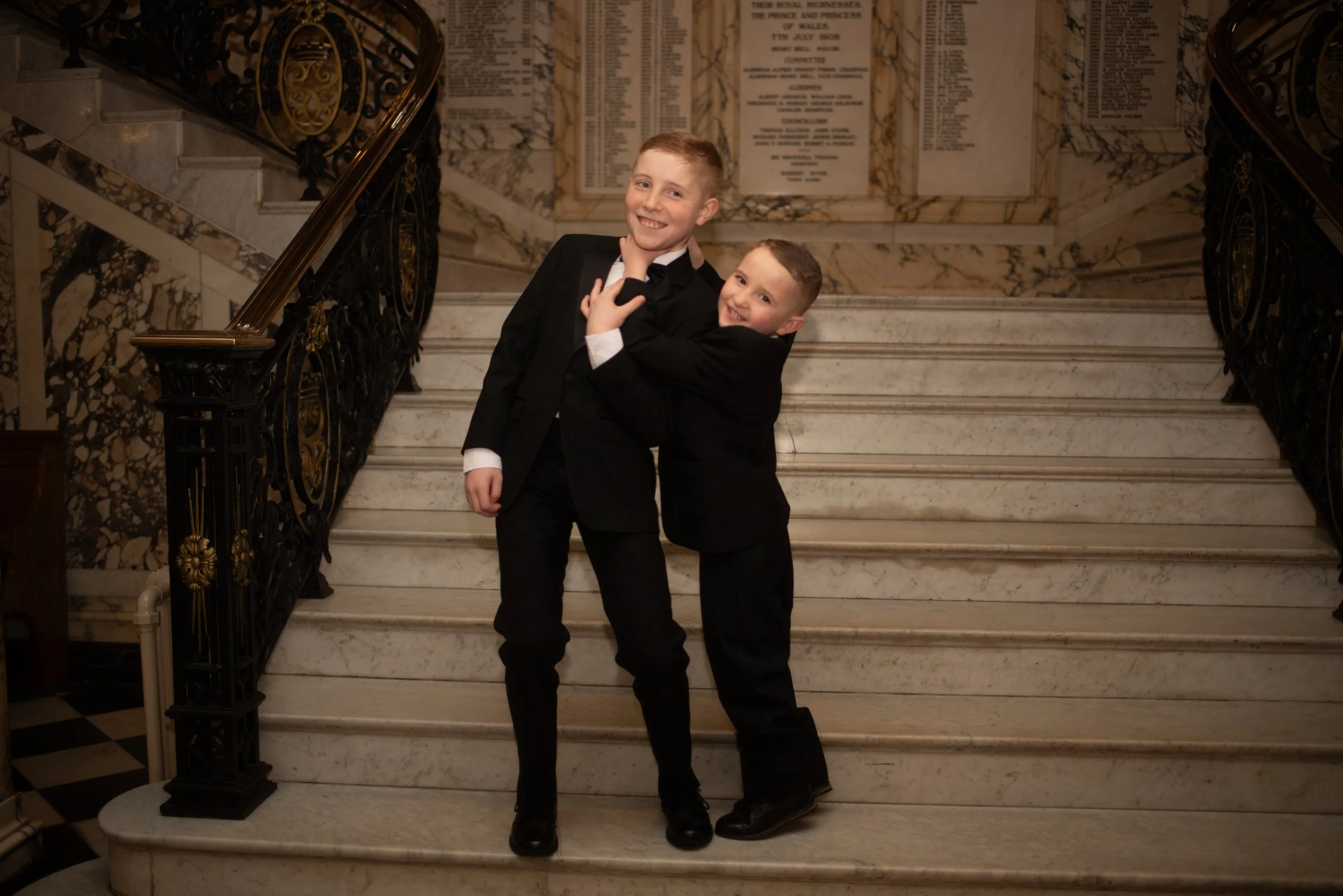 Two boys in formal tuxedos standing on a marble staircase, smiling and hugging each other.