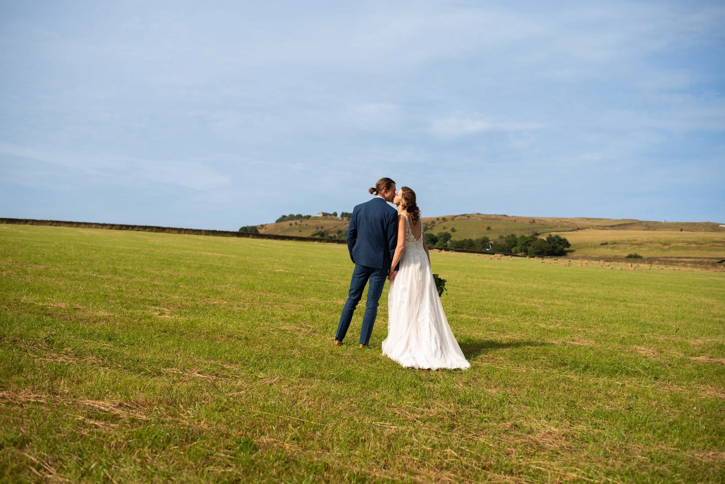 A bride and groom standing in an open grassy field, holding hands and kissing, with rolling hills in the background under a partly cloudy sky.