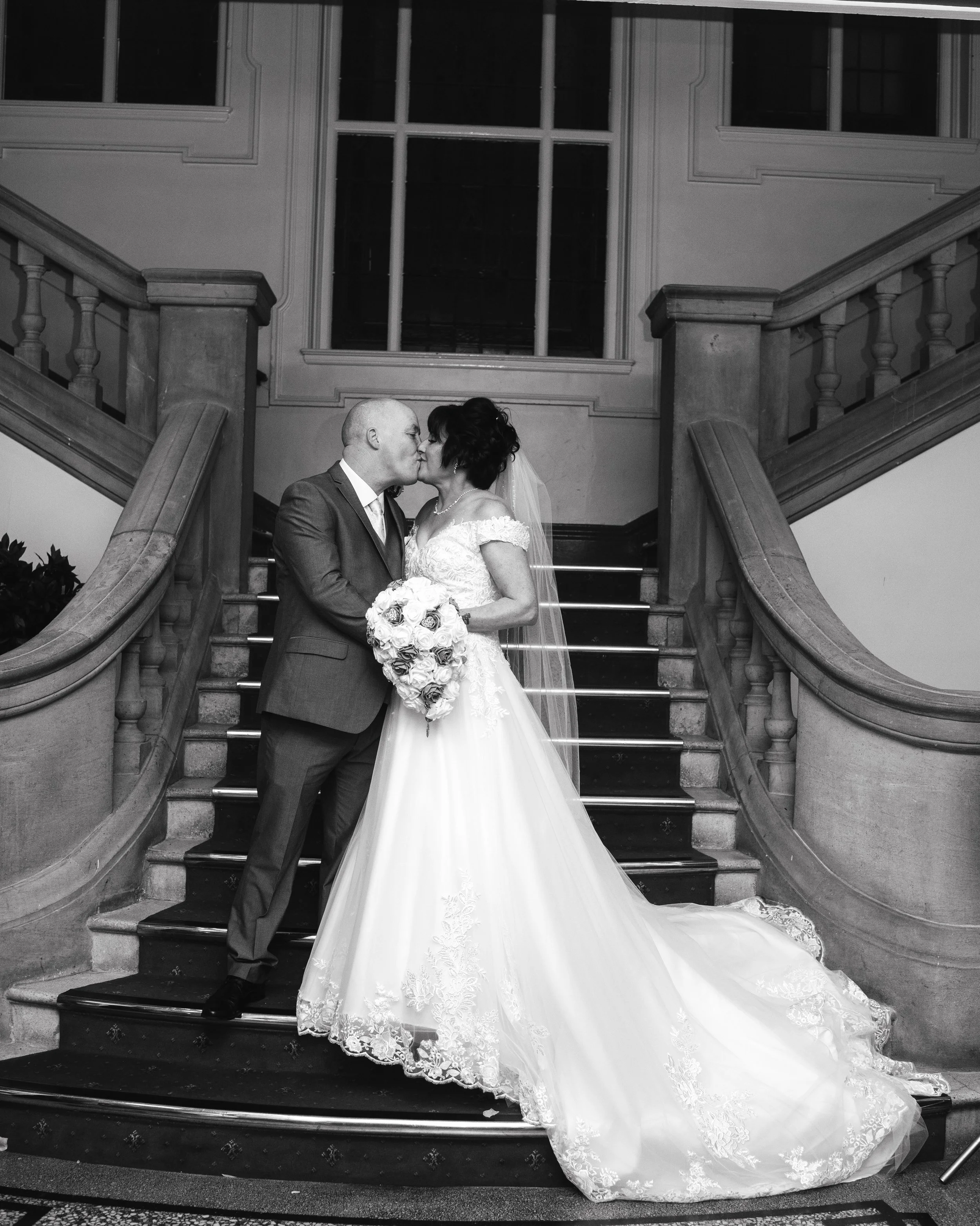 A bride and groom sharing a kiss on a staircase inside a building, with the bride holding a bouquet of flowers, in black and white.