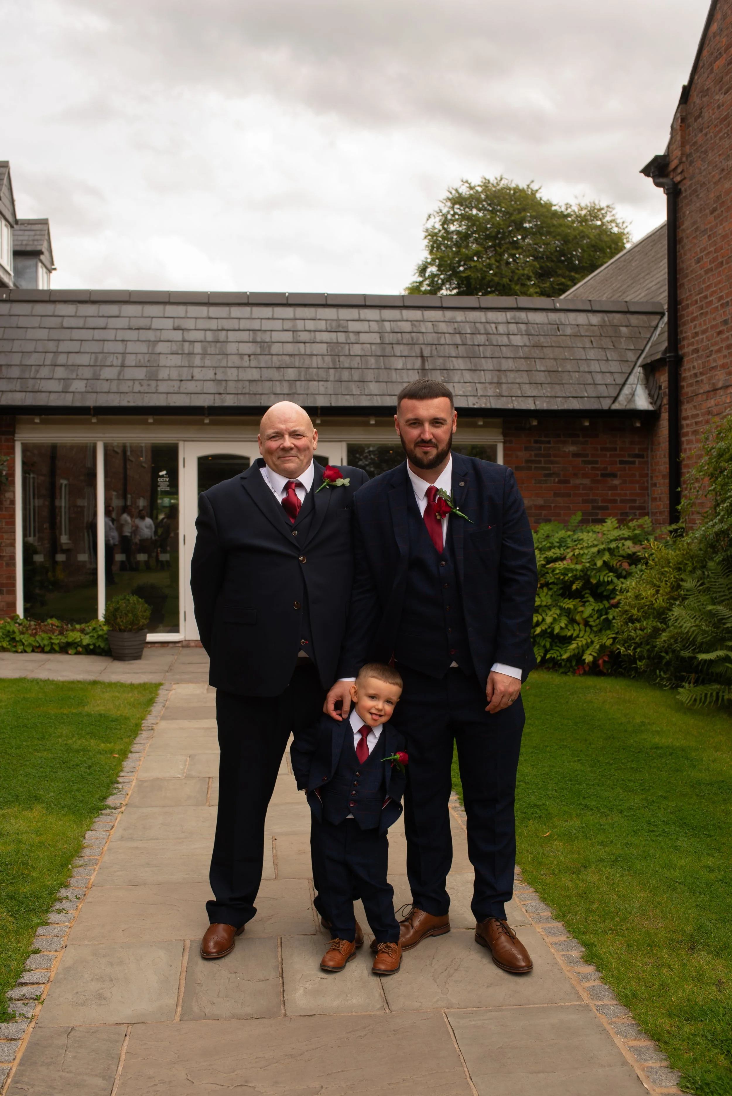 Three males in suits standing on a walkway outside a brick building, with the older man and younger man wearing boutonnières, and the young boy smiling. All appear dressed for a formal event.