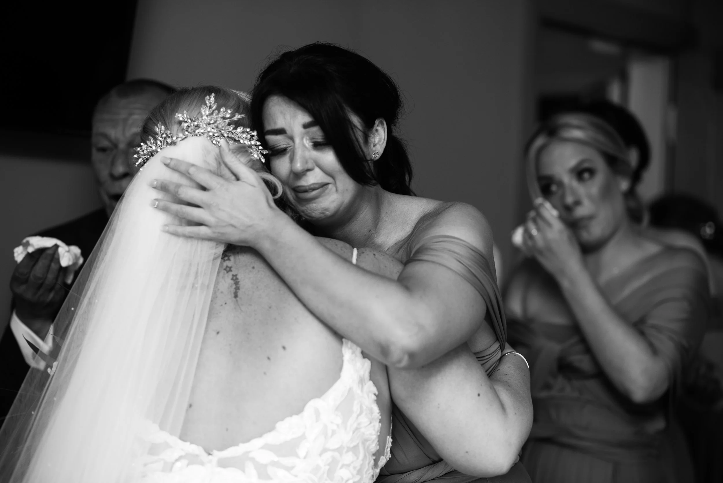 A woman embraces a bride in a heartfelt hug at a wedding, with tears on her face and emotional expressions. A man and another woman are in the background, also emotional.