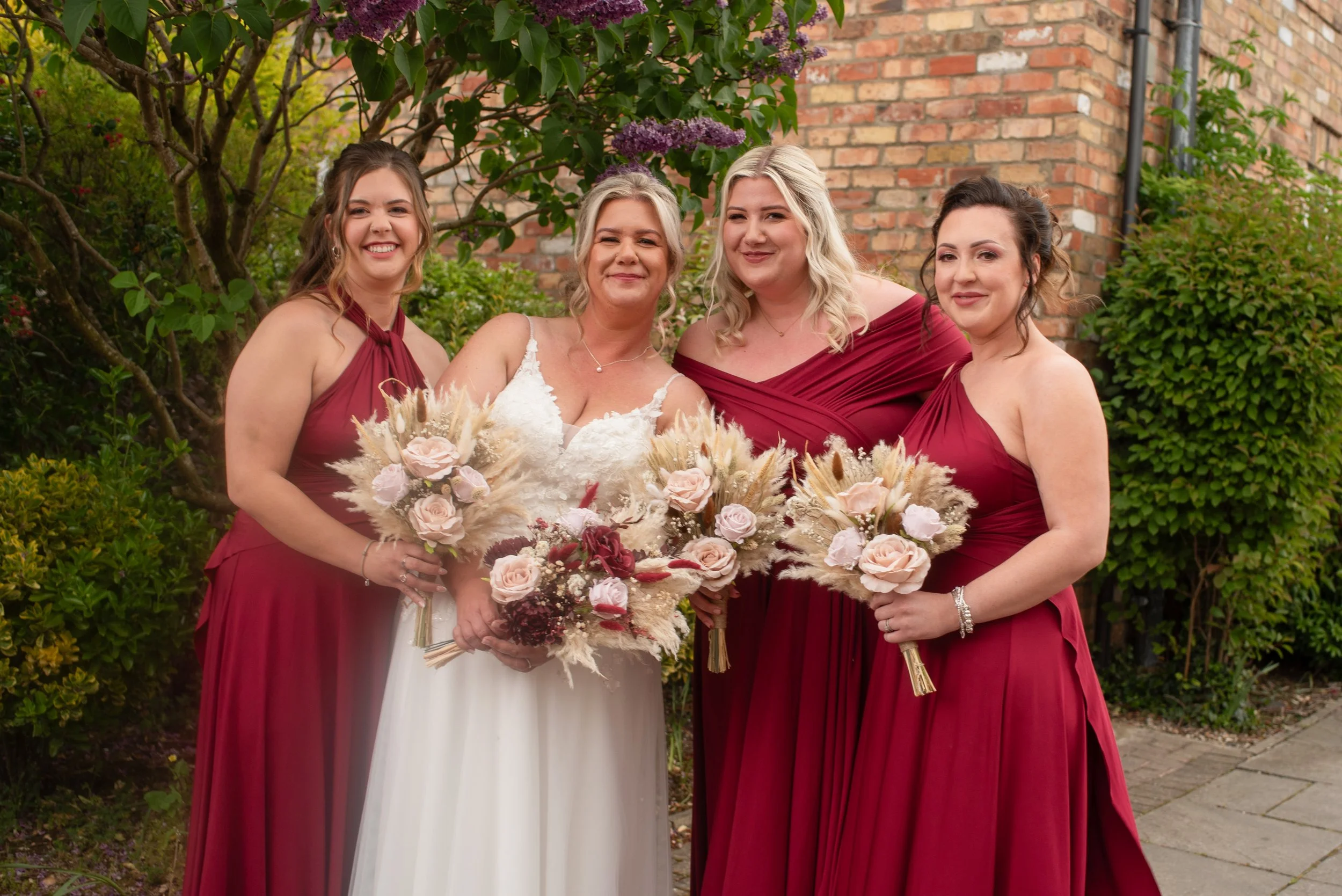 Four women standing outdoors, smiling and holding bouquets of flowers. One woman is wearing a white wedding dress, and the other three are in matching burgundy dresses. They are in front of green bushes and a brick wall.