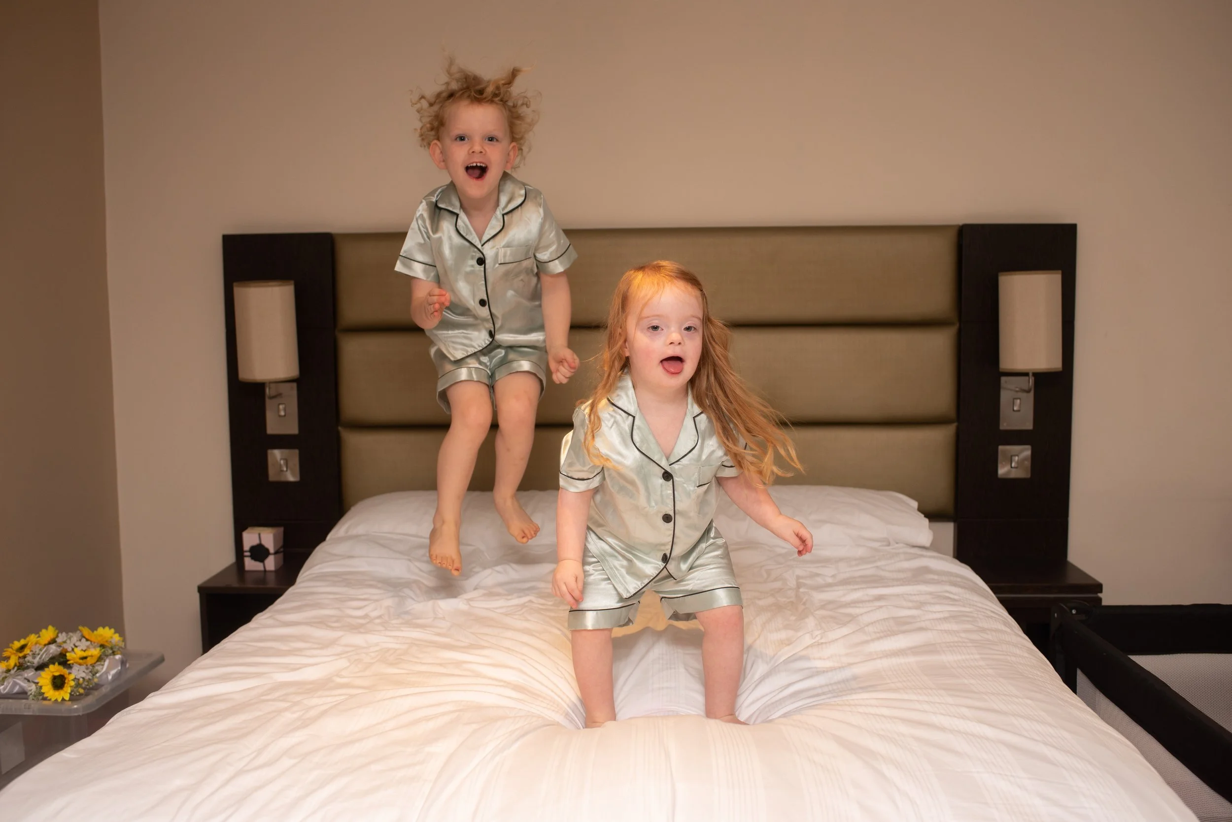 Two young girls in matching shiny pajamas jumping on a bed in a hotel room