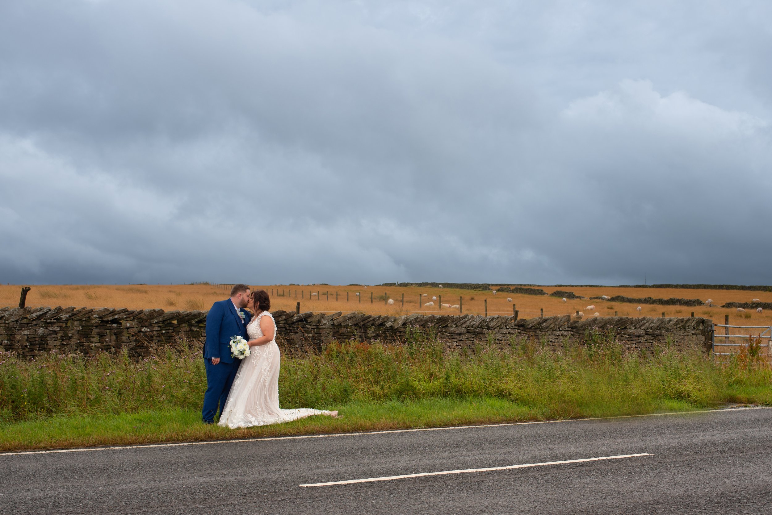 A bride and groom standing close to each other on the side of a rural road, with a stone wall and open fields with grazing sheep in the background, under a cloudy sky.