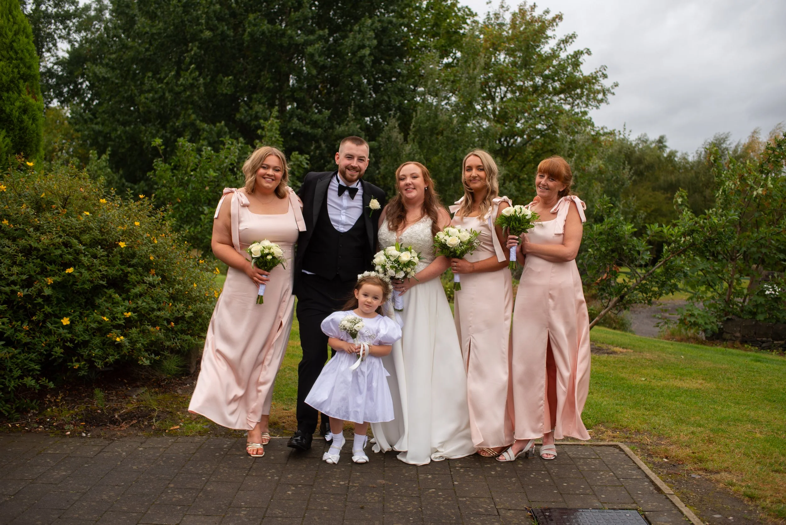 A wedding party of six adults and one child posing outdoors in a garden. The bride is in a white wedding dress, and the groom is in a black tuxedo. The women are in light pink dresses, holding bouquets. The young girl is in a white dress, holding a b