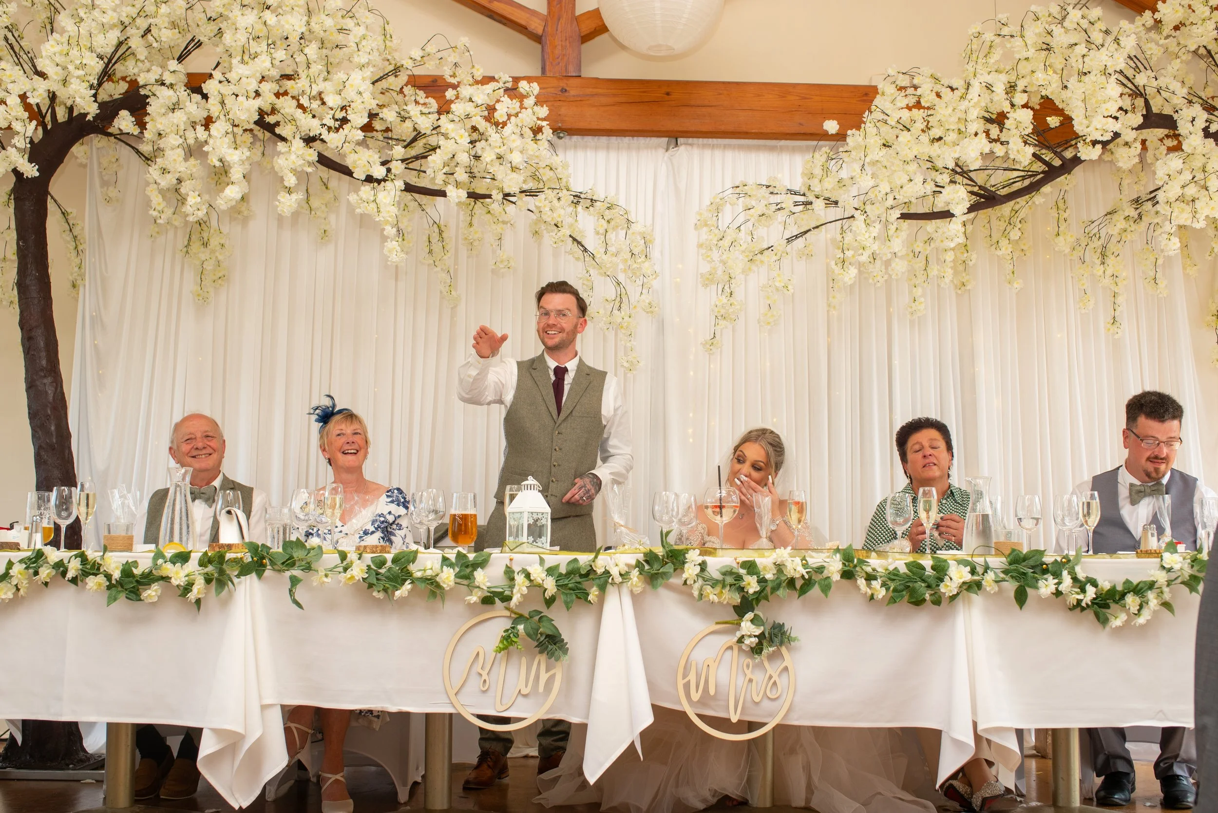 A wedding reception with a head table decorated with white flowers and greenery, under a canopy of white cherry blossoms. The bride and groom sit at the table with five guests, with a man standing and speaking or giving a toast. The setting is indoor
