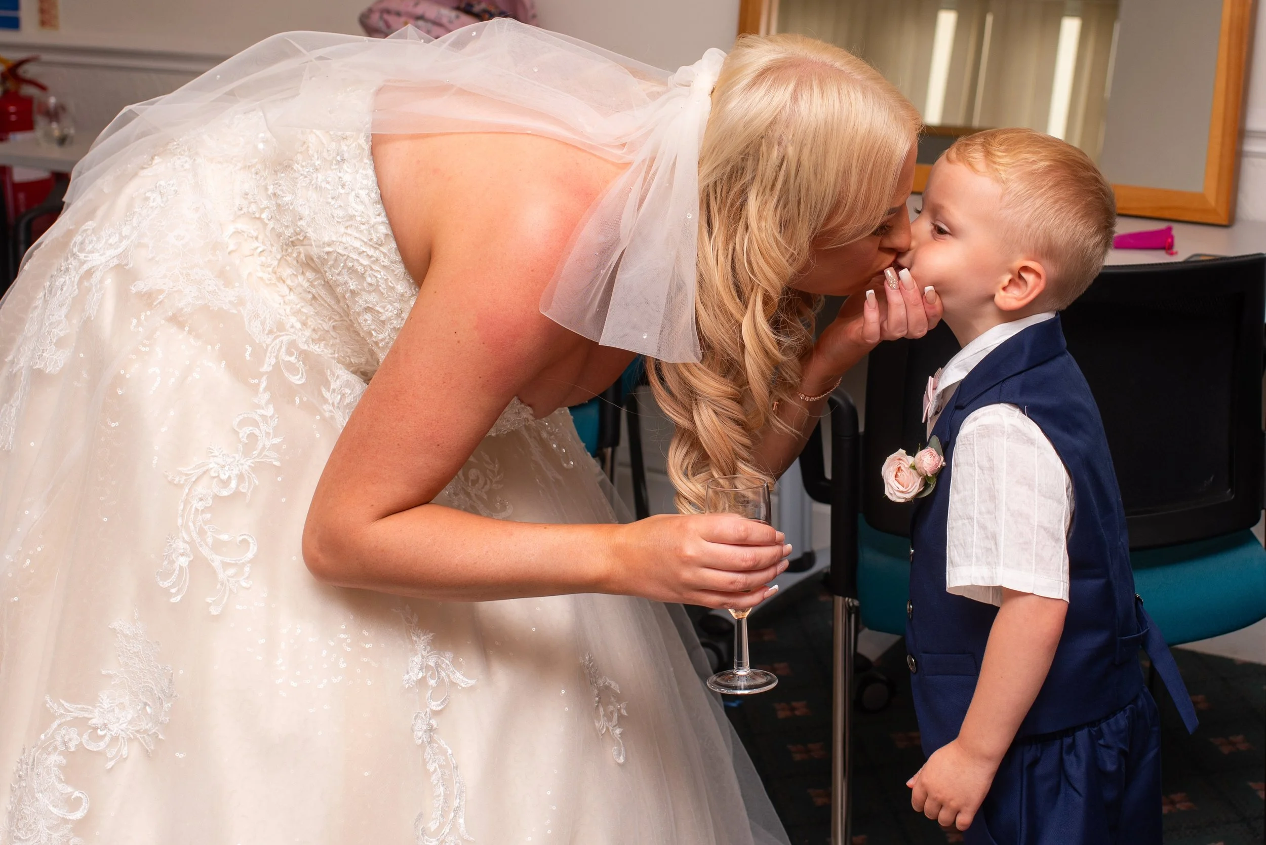 A woman dressed in a wedding gown leans over, kissing a young boy in formal attire, holding a glass of champagne, on a black chair with a green seat.