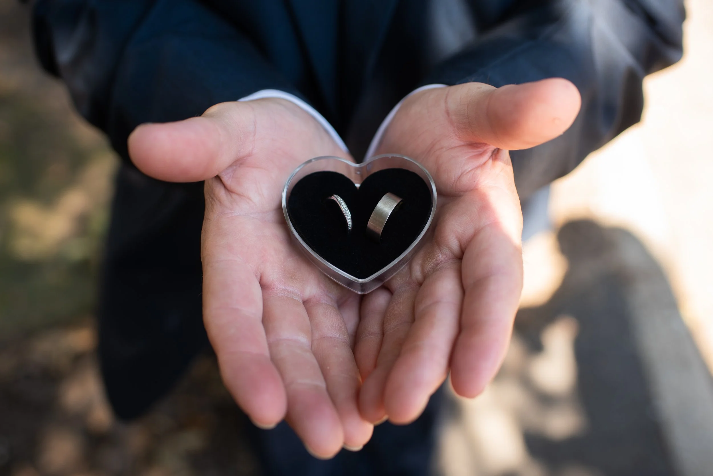 A person in a dark suit holding an open heart-shaped ring box with a wedding ring and a wedding band inside.