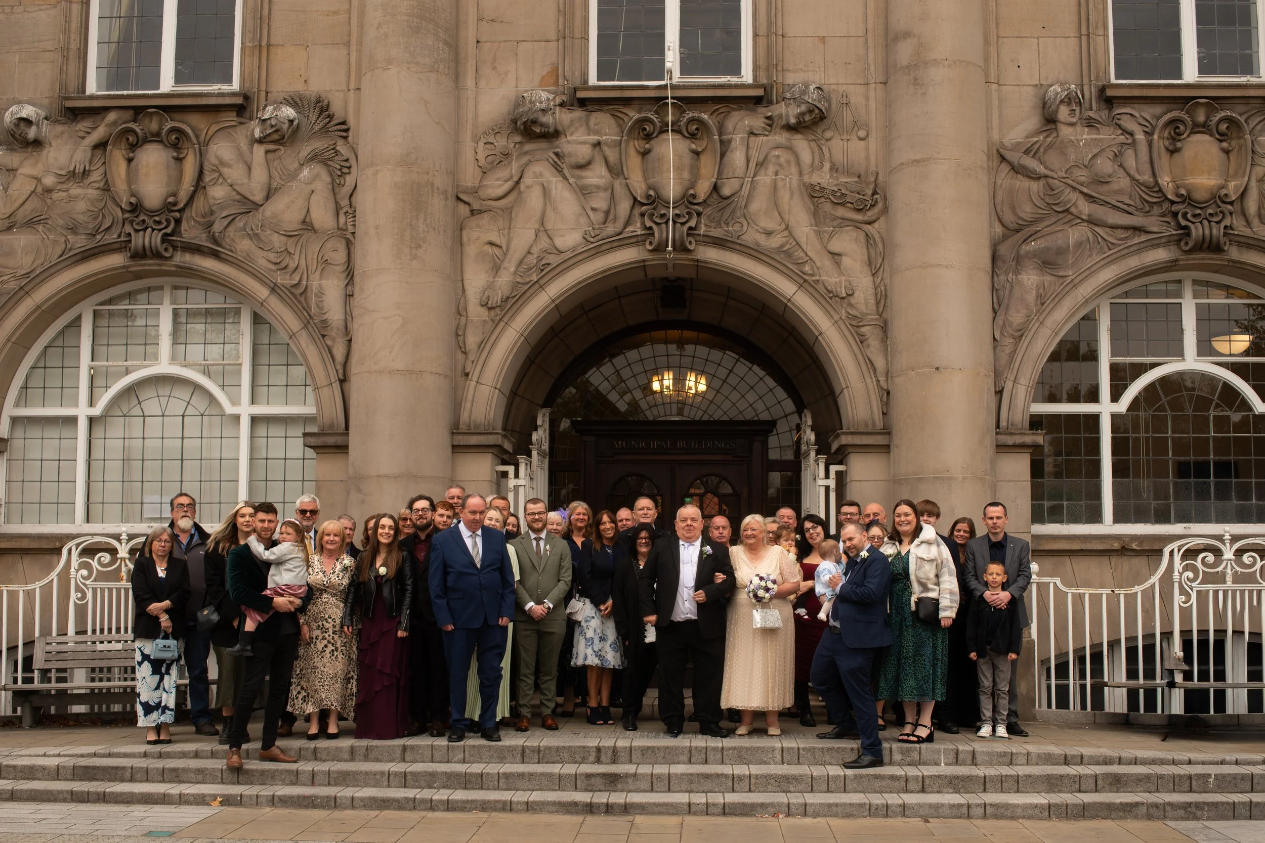 A large group of people dressed in formal attire standing in front of a historic building with stone carvings and large windows, posing for a group photo on steps.