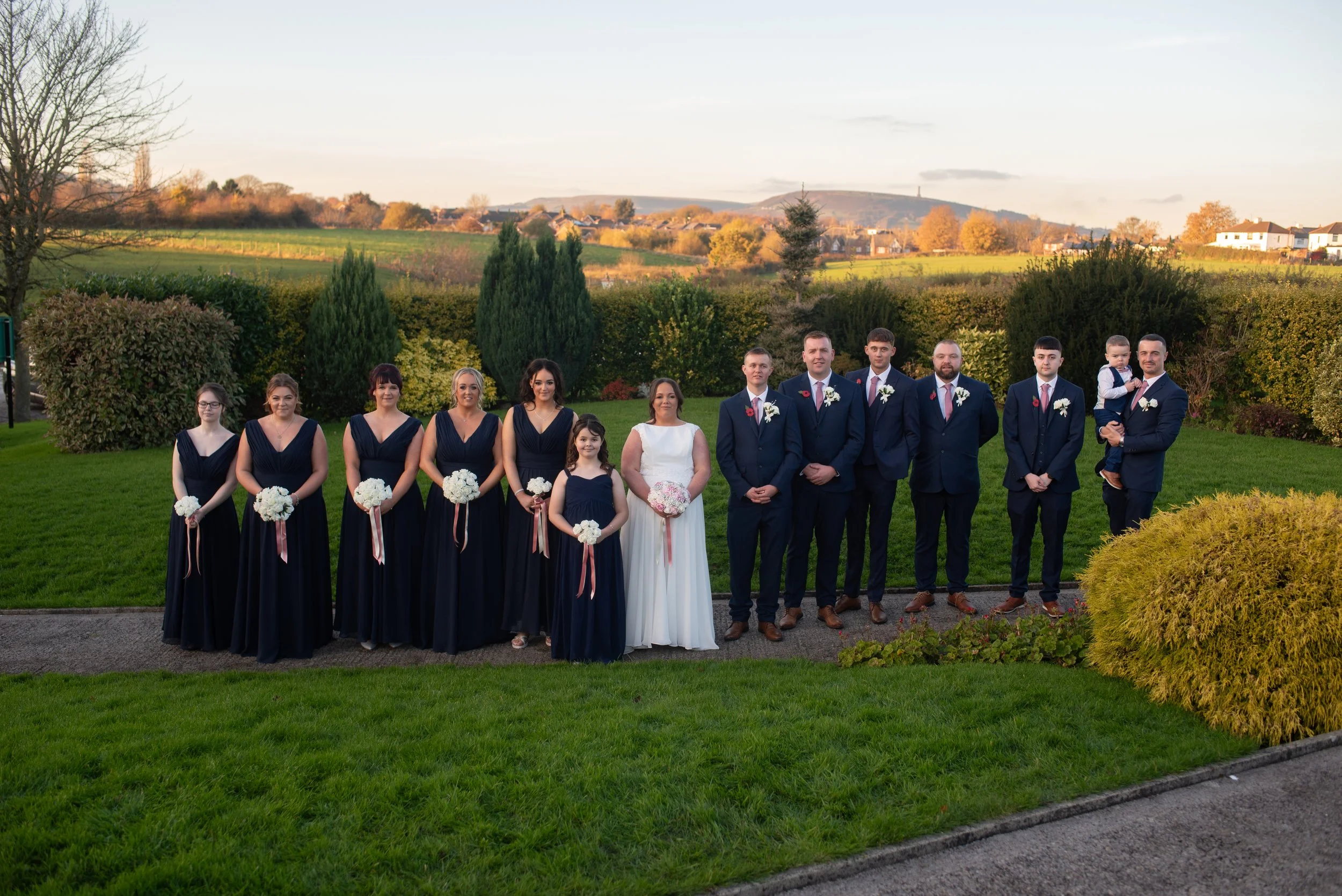 A wedding party outdoors with 11 people, including bridesmaids and groomsmen, standing on a pathway with a lush green background and hills in the distance, under a clear sky.