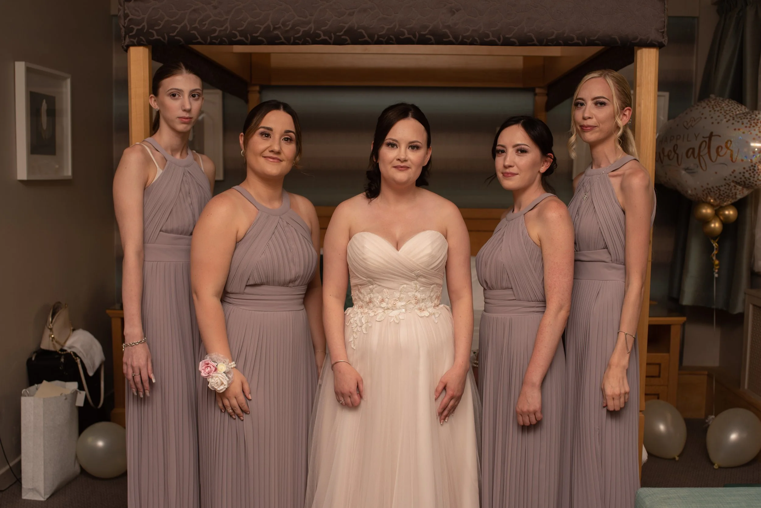 A bride in a white wedding gown standing with five bridesmaids in matching lavender dresses in an indoor setting decorated with balloons and framed photographs.