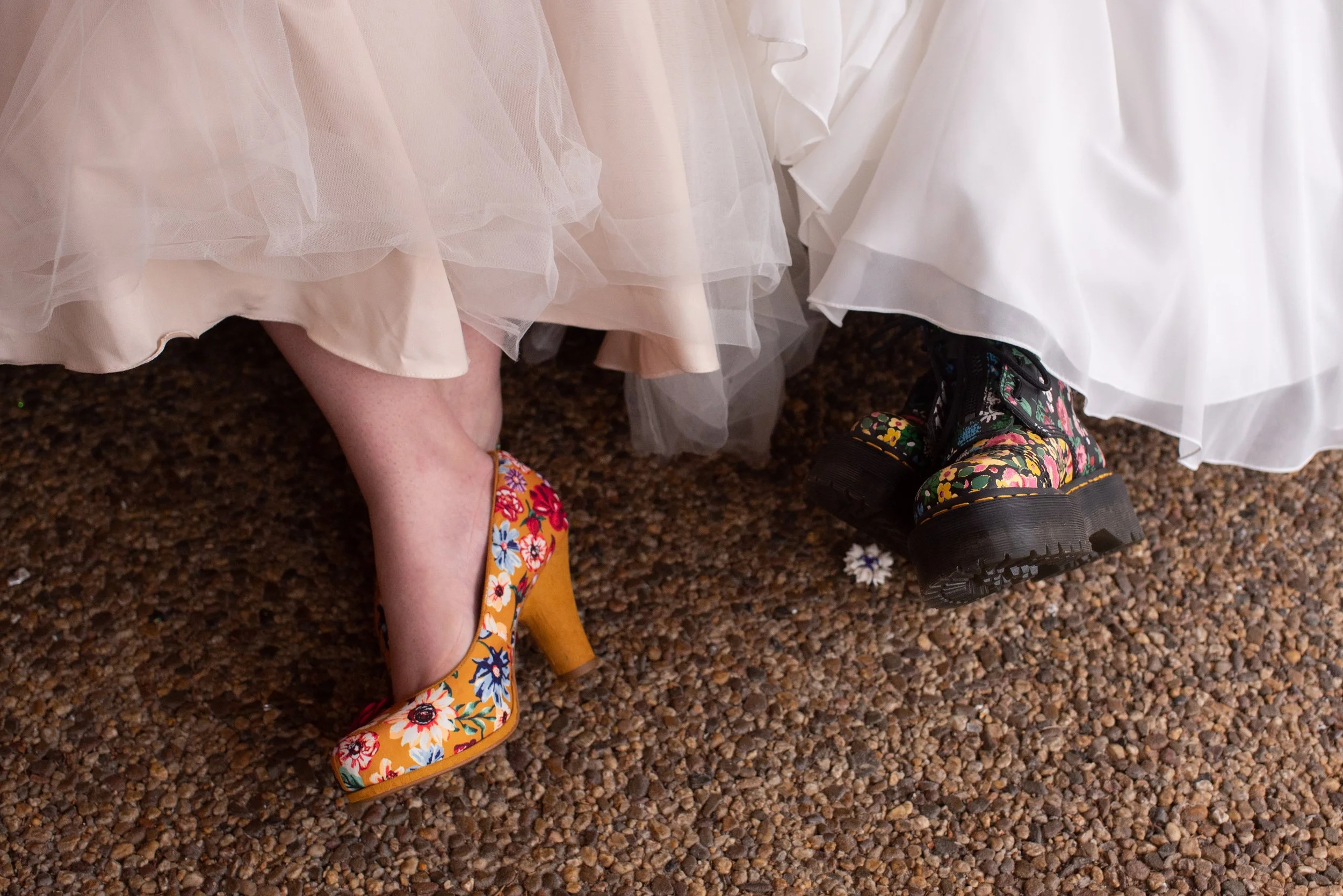 Two women in wedding dresses are wearing mismatched shoes: one in floral high heels, the other in chunky black boots with a floral pattern, standing on a pebble-covered ground.