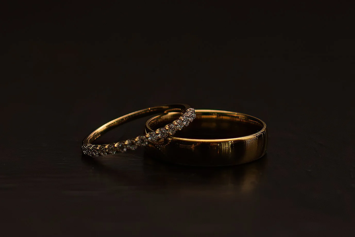 Close-up of gold wedding rings and a diamond-studded bracelet on a black surface, with a dark background.