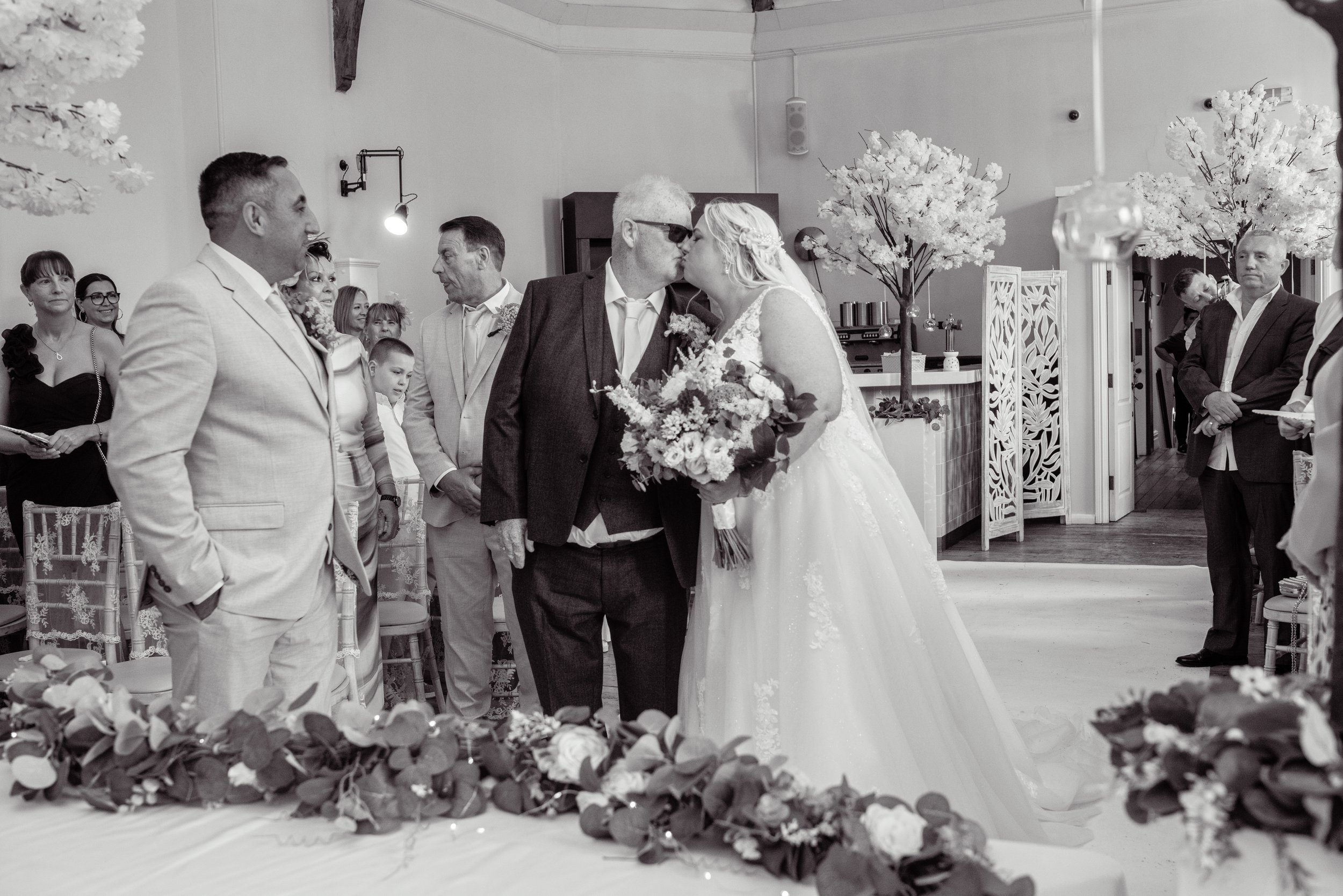 A bride and an older man, possibly her father, sharing a kiss during a wedding ceremony. The bride is holding a bouquet of flowers and wearing a wedding gown, while the man is dressed in a suit jacket and sunglasses. Several guests are watching, some