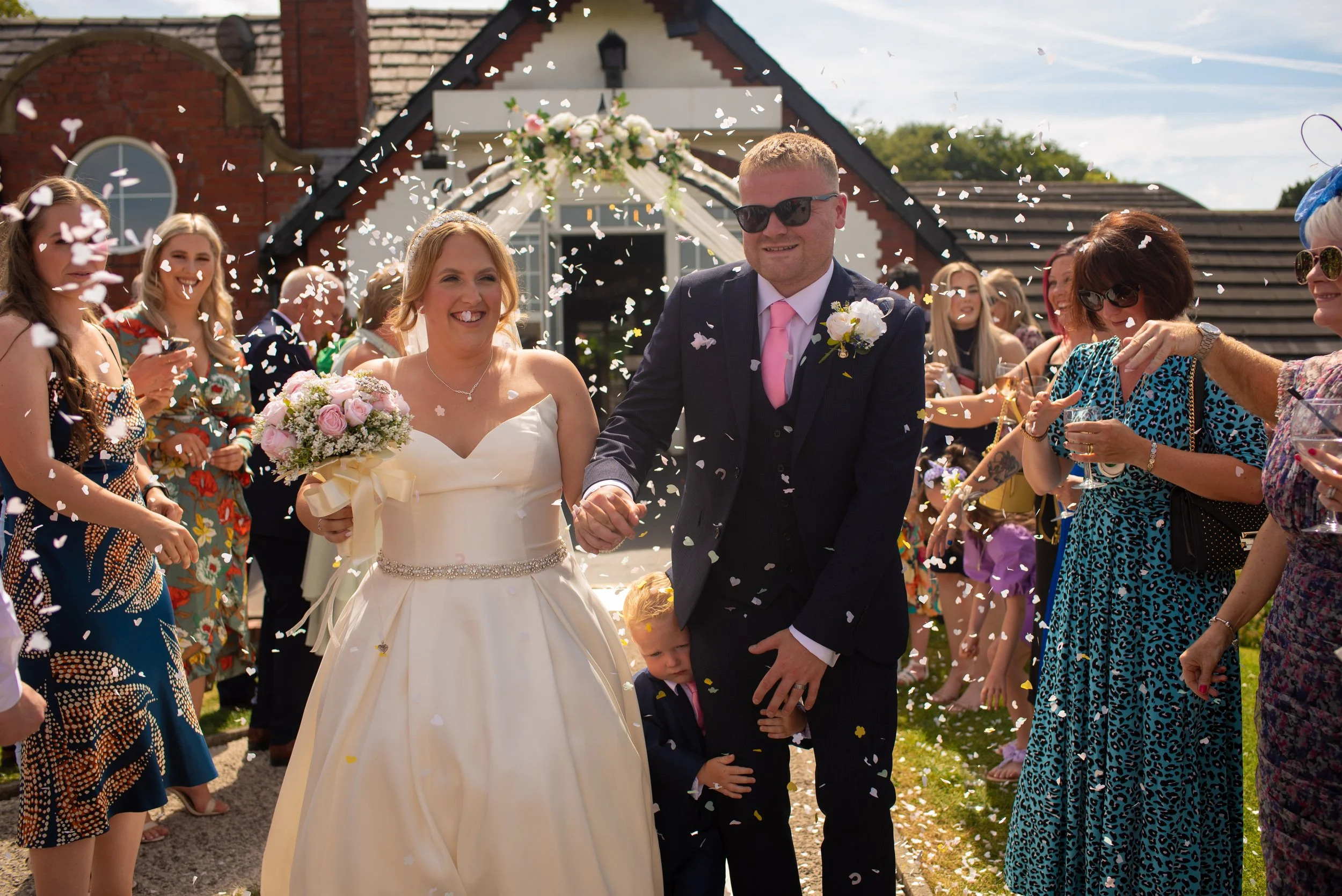 A bride and groom walking hand in hand after their wedding ceremony, surrounded by friends and family throwing confetti outside a decorated building.
