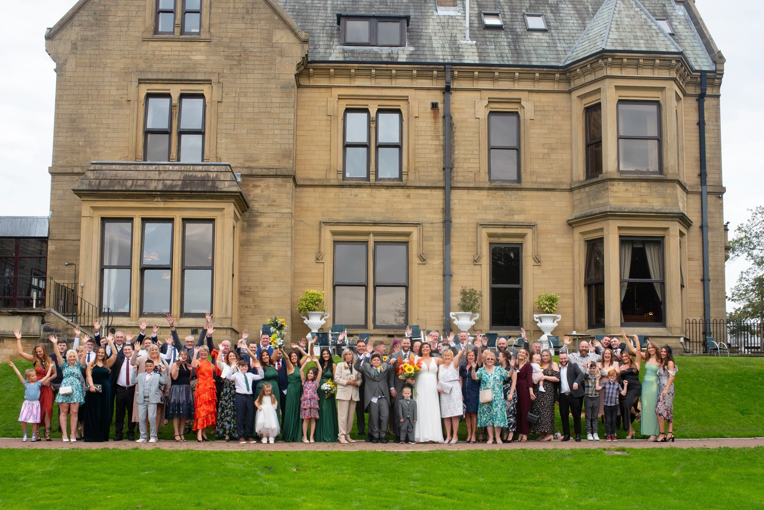 Group of people, including a bride and groom, cheering and waving in front of a large old stone building with multiple windows and decorative planters on the balcony.