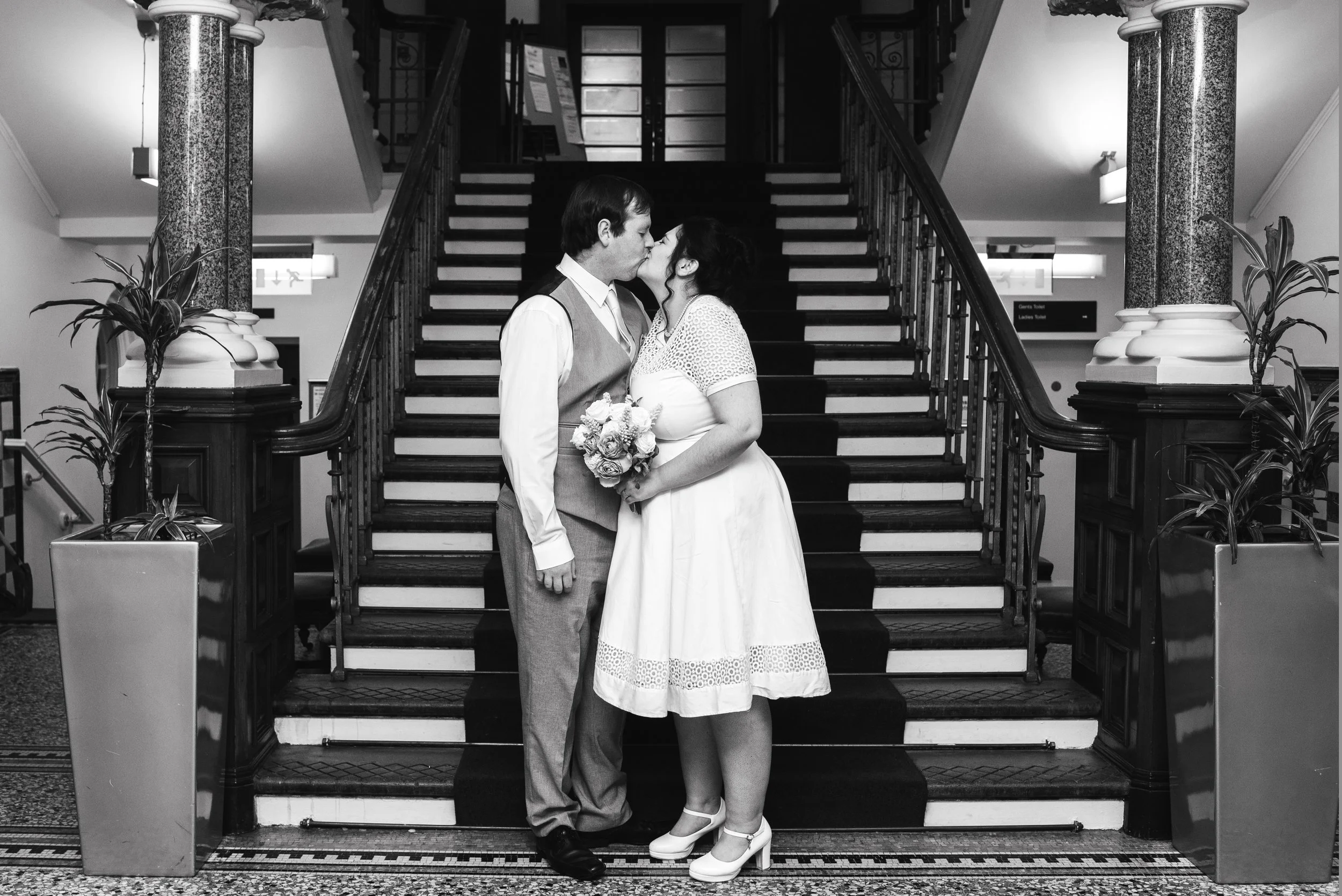 Black and white photo of a couple in vintage wedding attire gently touching foreheads and kissing on a staircase inside a building, with potted plants on either side.