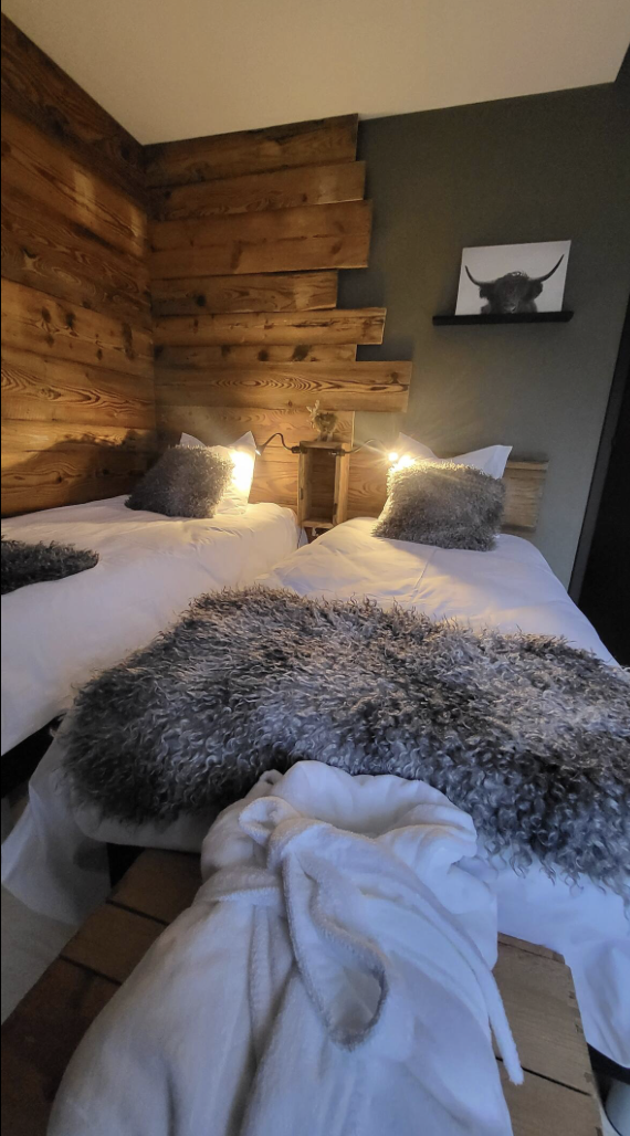 A cozy bedroom with two beds featuring white bedding, gray furry pillows, and blankets. The headboards are wooden, with a wall made of wood planks and a dark painted section. There is a small shelf with a black and white picture of an animal, possibly a buffalo or bison, above the beds. Warm lighting adds to the cozy ambiance.