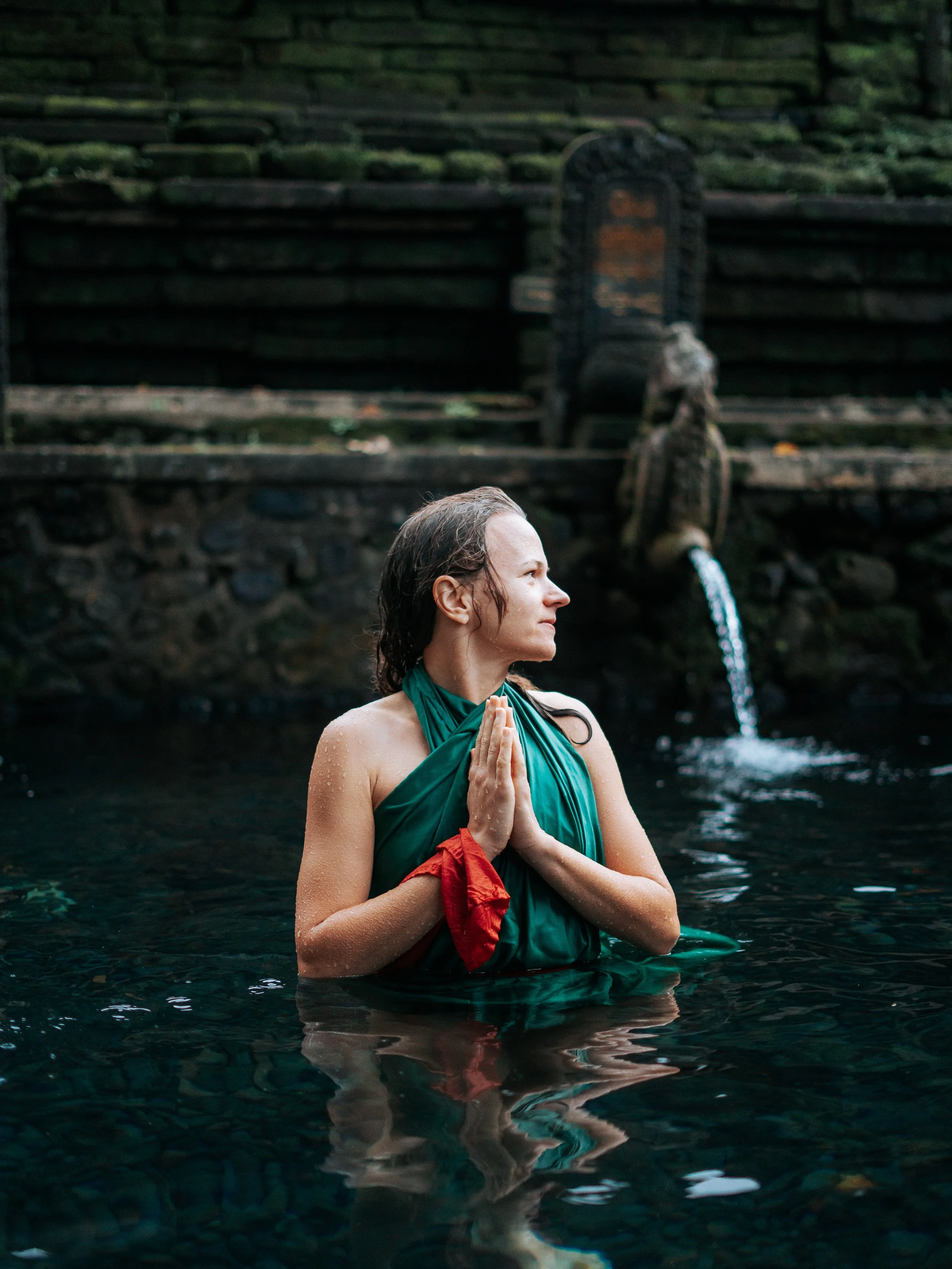 A woman standing in a pool of water, wearing a green cloth tied around her body, with her hands in prayer, and wet hair. In the background, there is a stone water fountain with water flowing out.