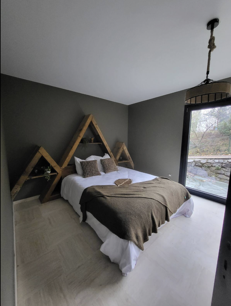 A bedroom with a dark gray wall, a bed with white linens and brown pillows, and a wooden geometric headboard. There's a large window to the right, showing a view of trees and rocks outside.