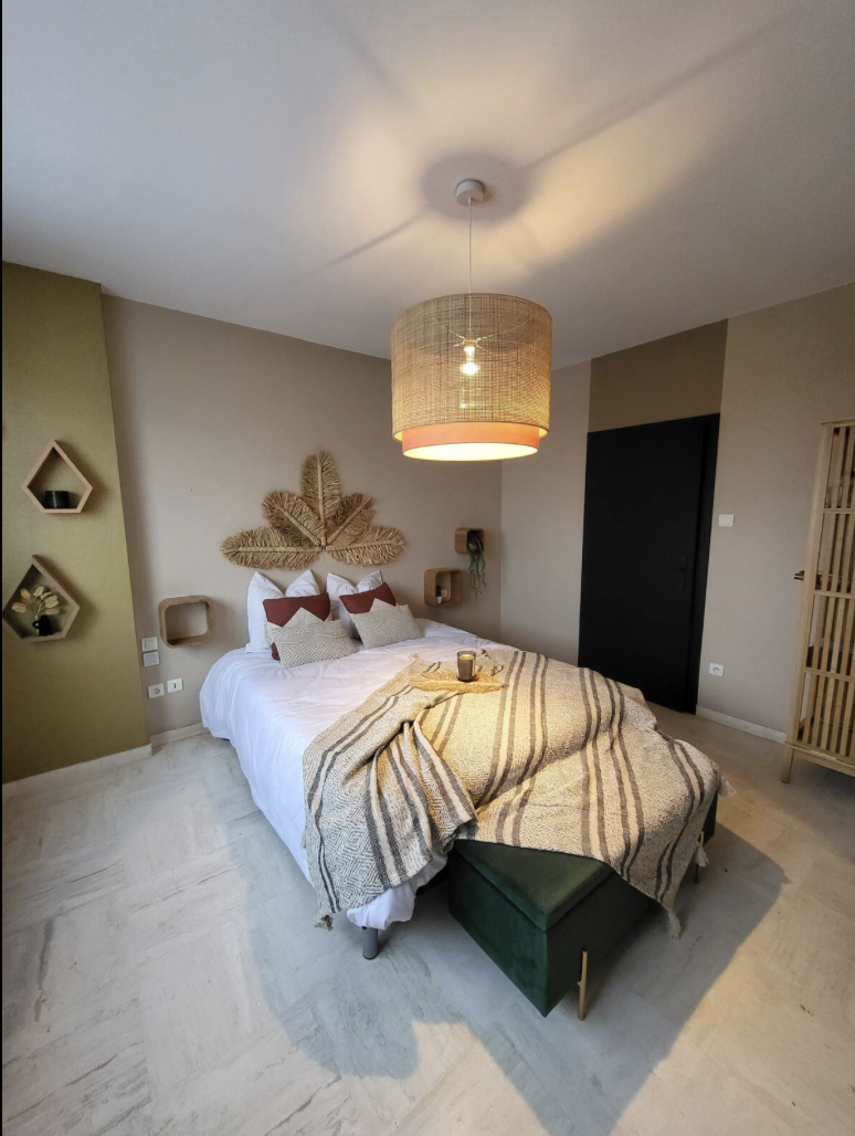 A cozy bedroom with a bed decorated with white and brown pillows, a woven wall art above the bed, a hanging ceiling light, and small wall shelves on a neutral-colored wall.