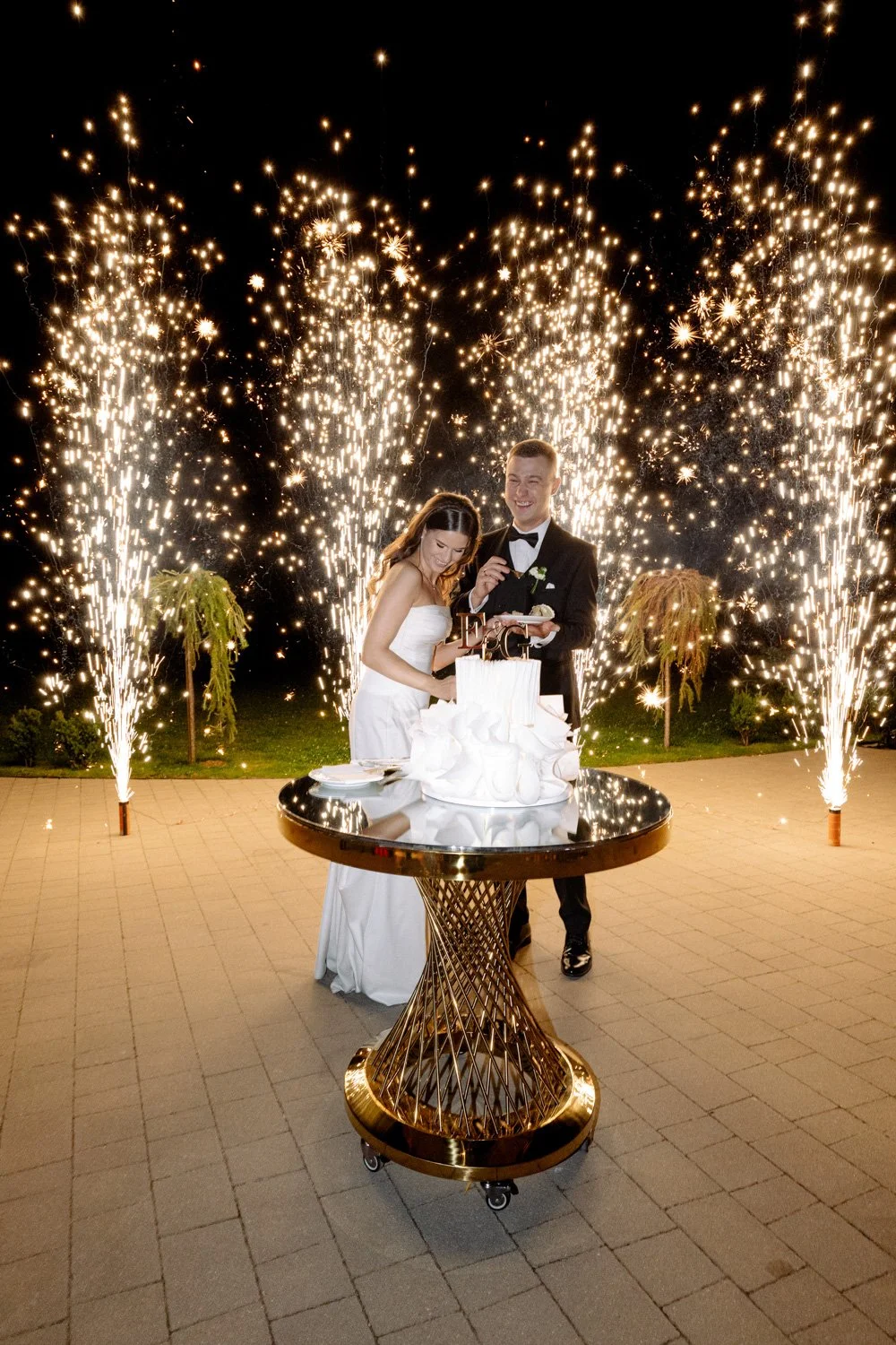 A newlywed couple in wedding attire cutting a wedding cake outdoors at night, surrounded by sparklers and fireworks.