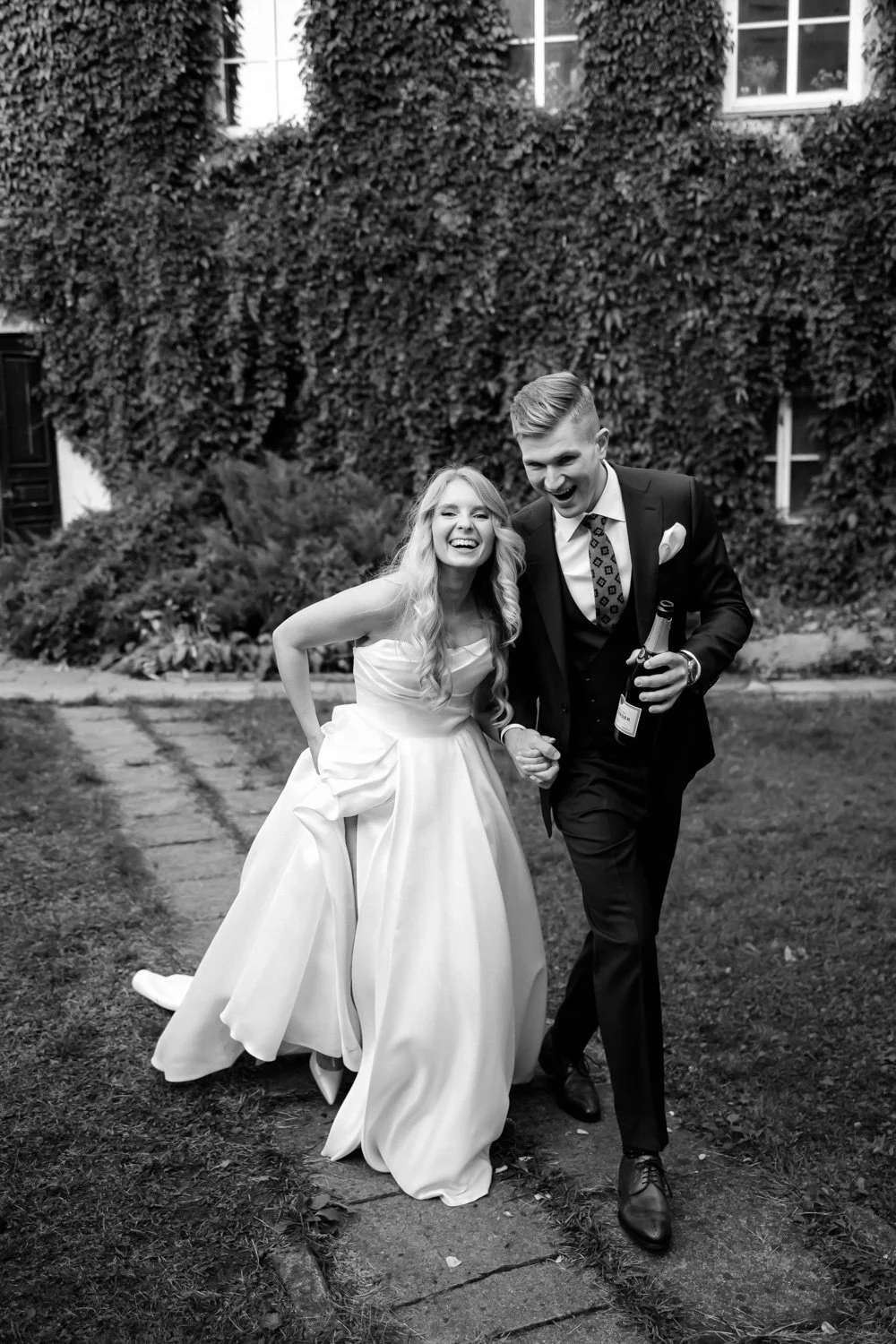 A bride and groom holding hands in front of a grand hotel entrance with yellow flowering trees and European flags.