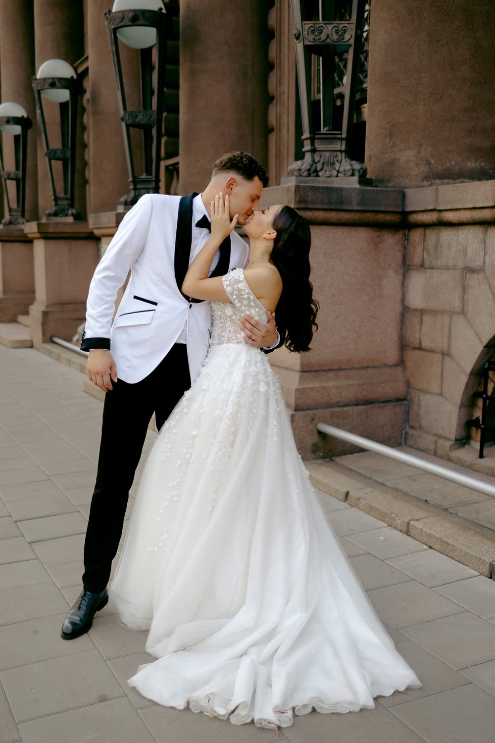 A bride and groom share a romantic moment outdoors. The groom wears a white tuxedo with black accents, and the bride is in a white off-the-shoulder wedding gown with lace details. They are standing close, touching foreheads and gazing into each other