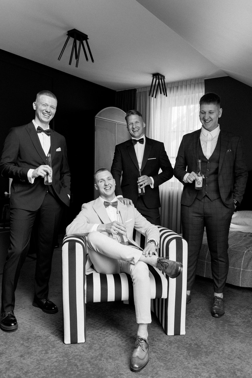 Five men dressed in formal suits, some with bow ties, gathering in a room with a striped armchair, smiling and holding drinks.