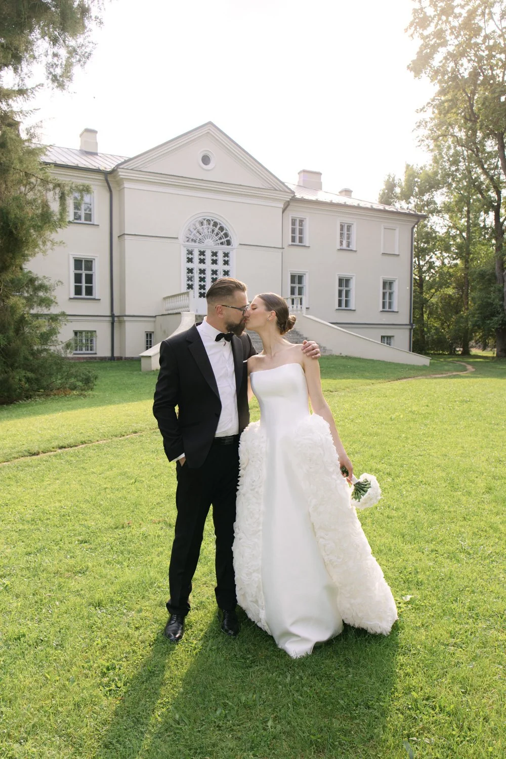 A bride and groom sharing a kiss outdoors in front of a white mansion, with the bride wearing a white strapless wedding gown and holding a bouquet, and the groom in a black tuxedo and bowtie.