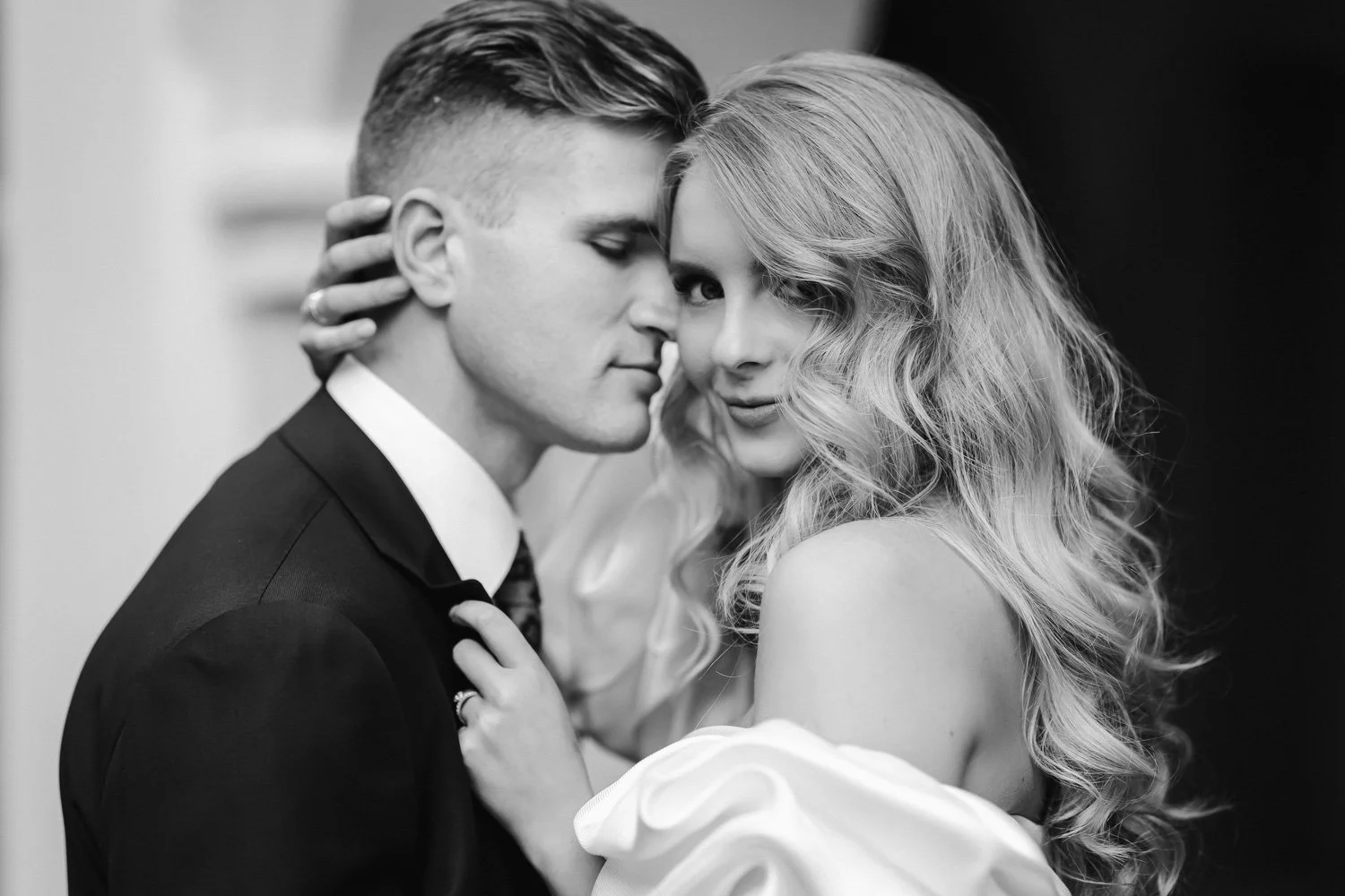 A black and white photo of a man and woman in an intimate pose, with their faces close together. The woman has wavy hair and is wearing an off-shoulder dress, while the man has short hair and is dressed in a suit. They appear to be in a romantic mome