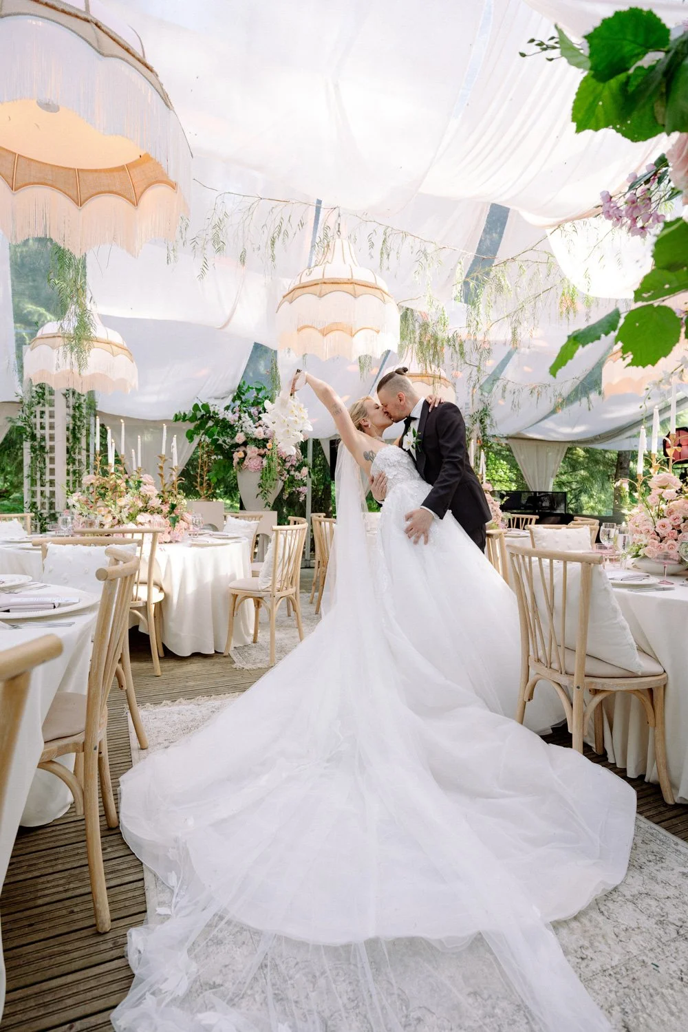 A bride and groom sharing a kiss at their wedding reception, with the bride in a white gown and the groom in a black tuxedo, surrounded by decorated tables and floral arrangements under a canopy of white fabric and hanging lace umbrellas.