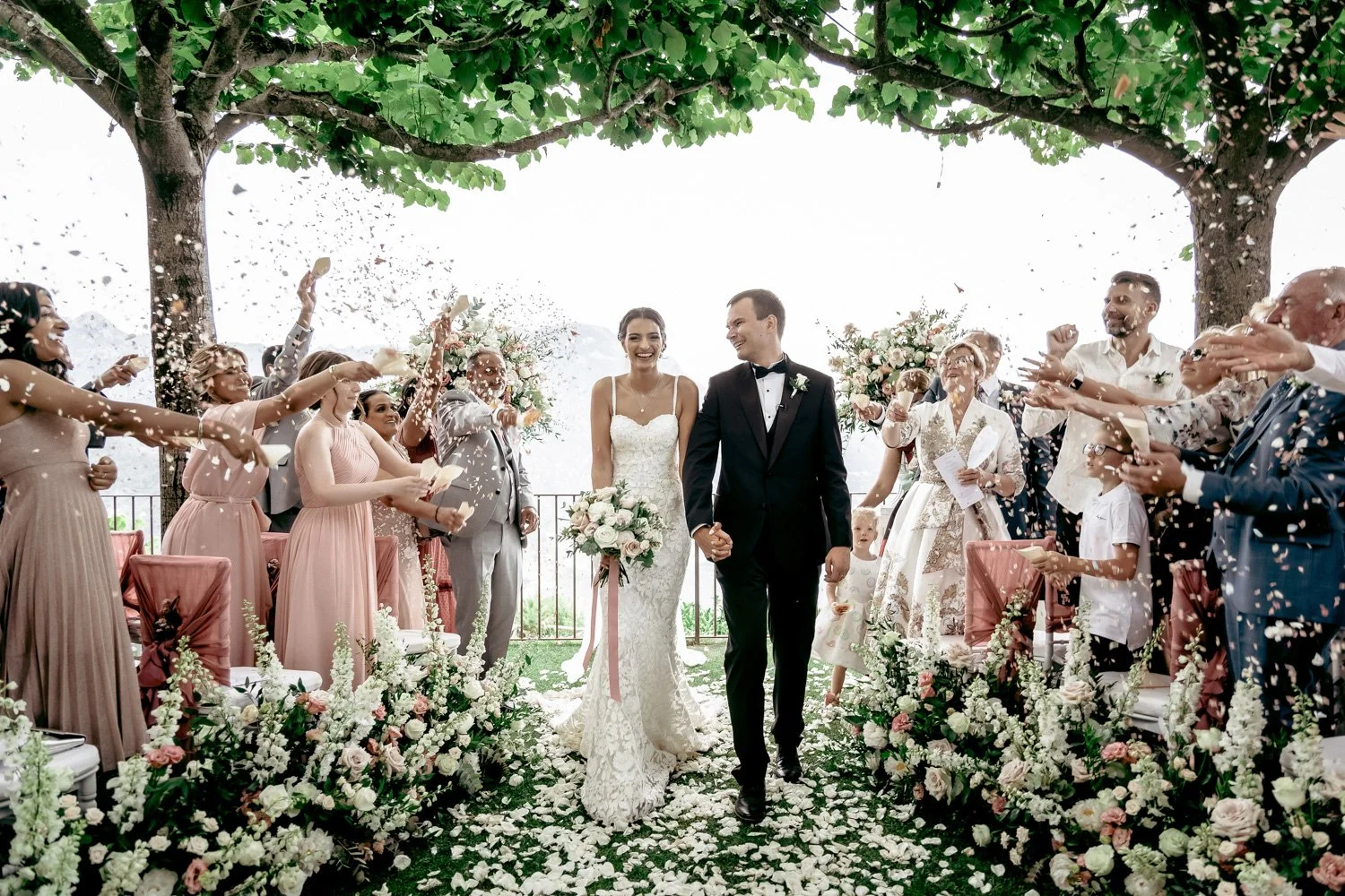 Bride and groom walking down the aisle at their wedding celebration, surrounded by guests throwing flower petals, under a green leafy canopy with floral arrangements along the aisle