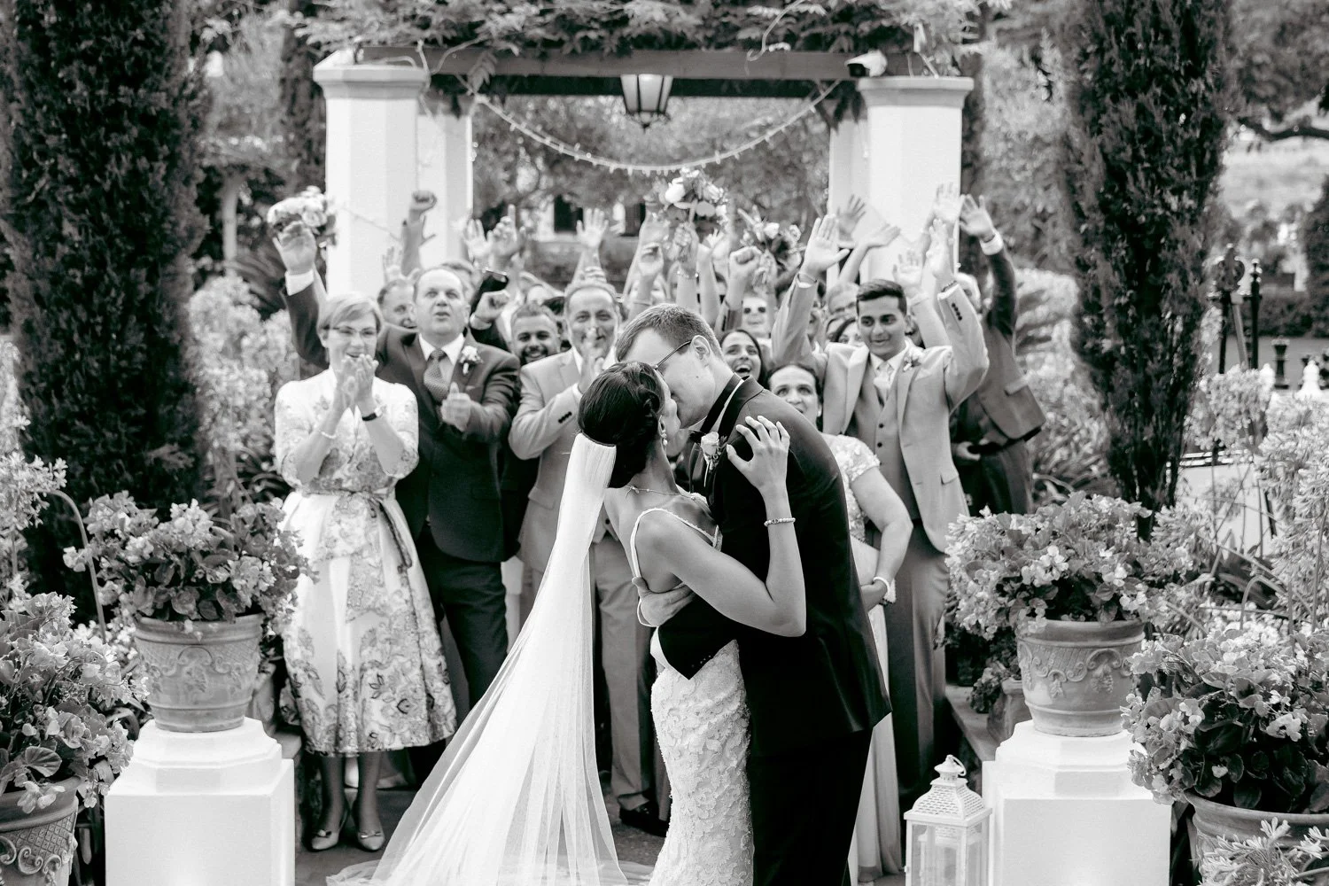 A black-and-white photo of a wedding celebration where a bride and groom share a kiss in front of a crowd of guests.